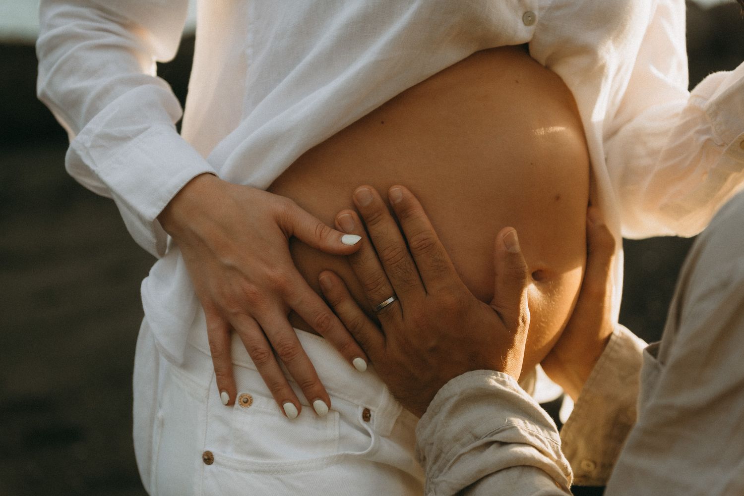 Couple tenderly cradling a pregnant belly, both wearing wedding rings, dressed in white clothing.