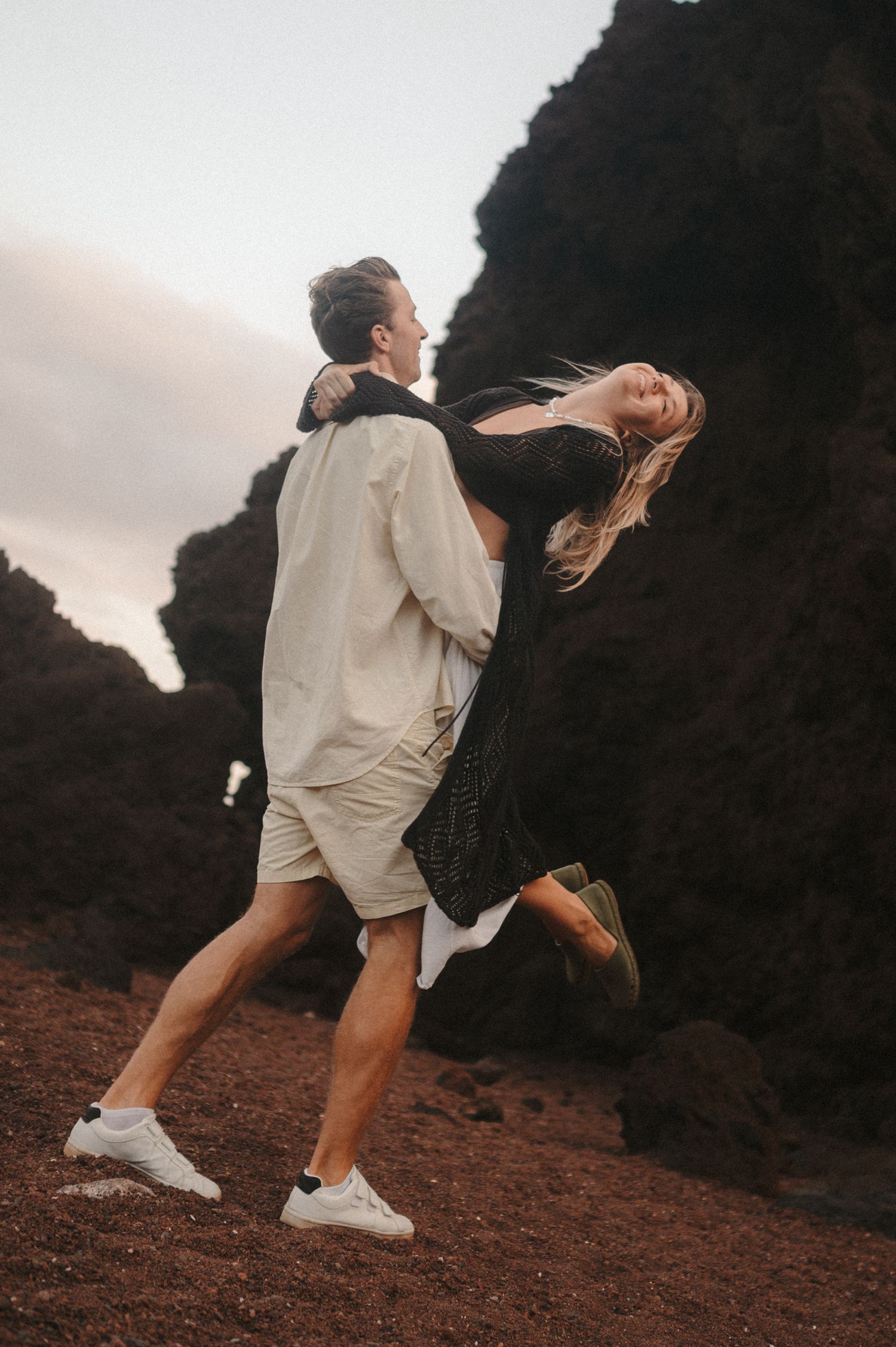 Young romantic couple under Montaña Roja, he is holding her in his arms while her hair flows in the wind, both smiling during a photoshoot i
