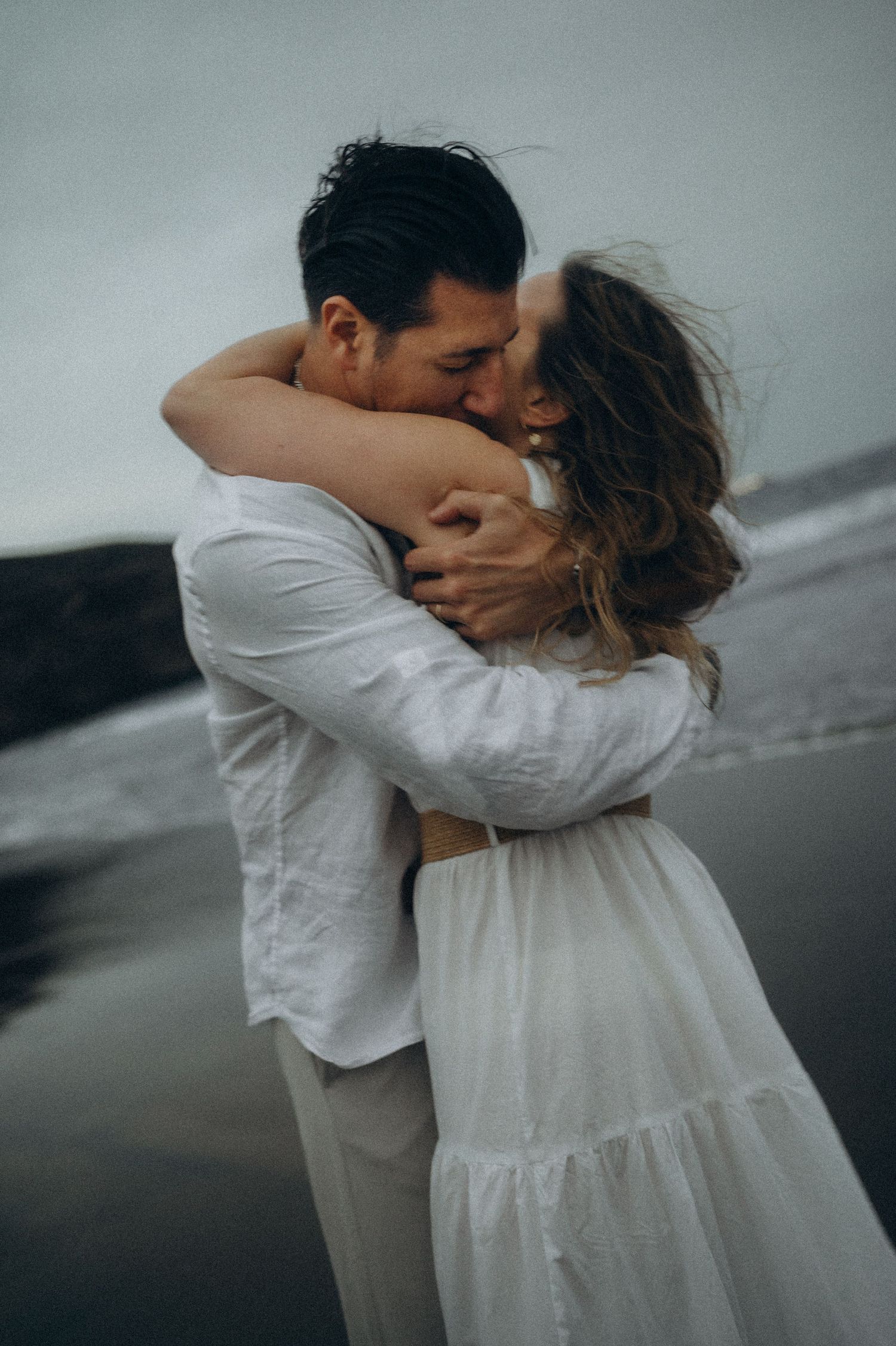 Couple embracing and kissing on a moody beach, both wearing white outfits, with wind blowing through her hair.