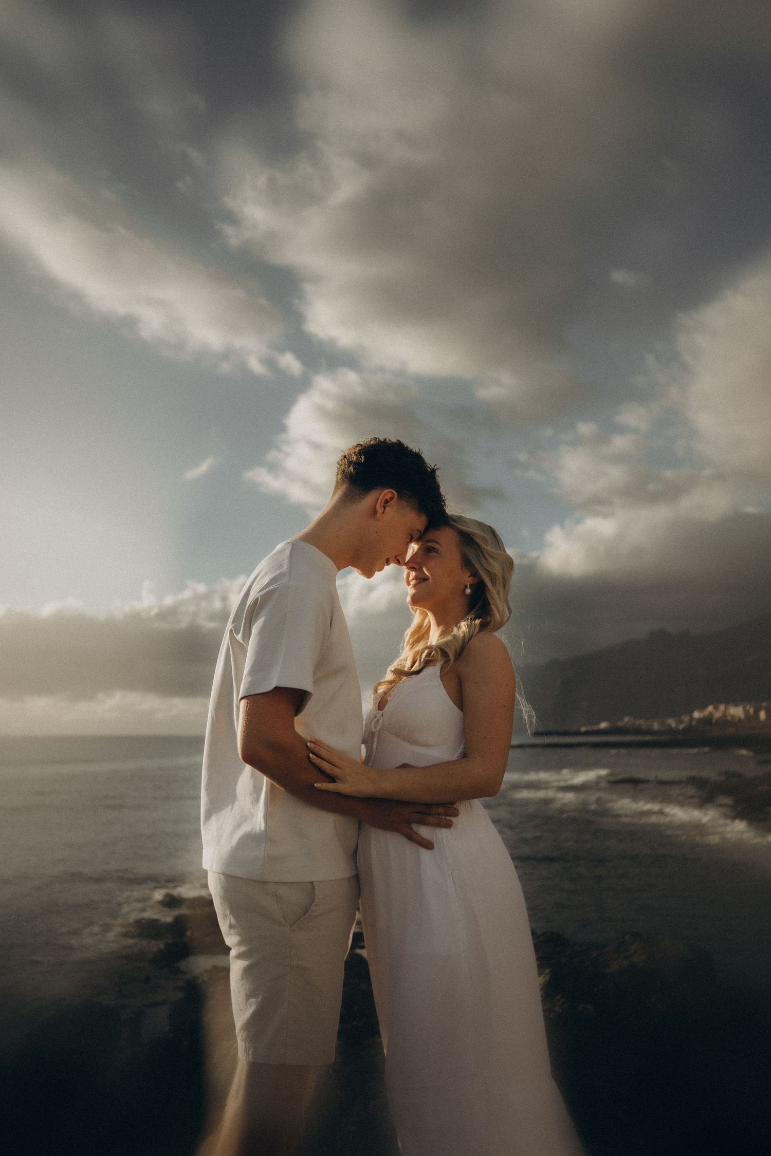 Couple in white outfits embracing near ocean with dramatic cloudy sky backdrop at golden hour.