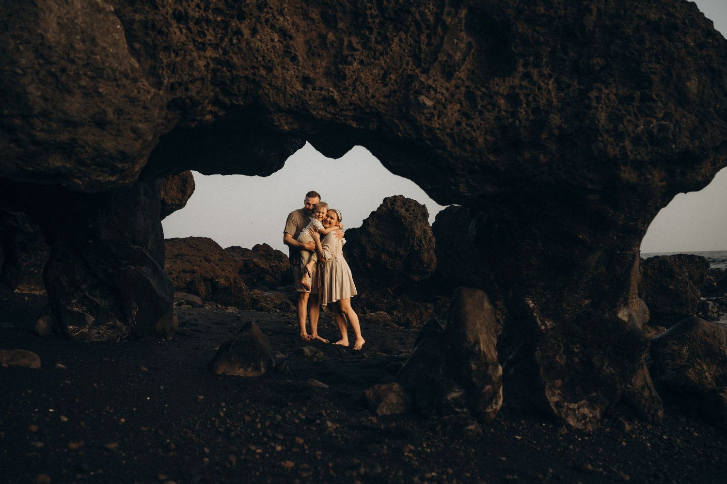 Couple embracing romantically framed by dramatic volcanic rock arch on a dark rocky beach at sunset.