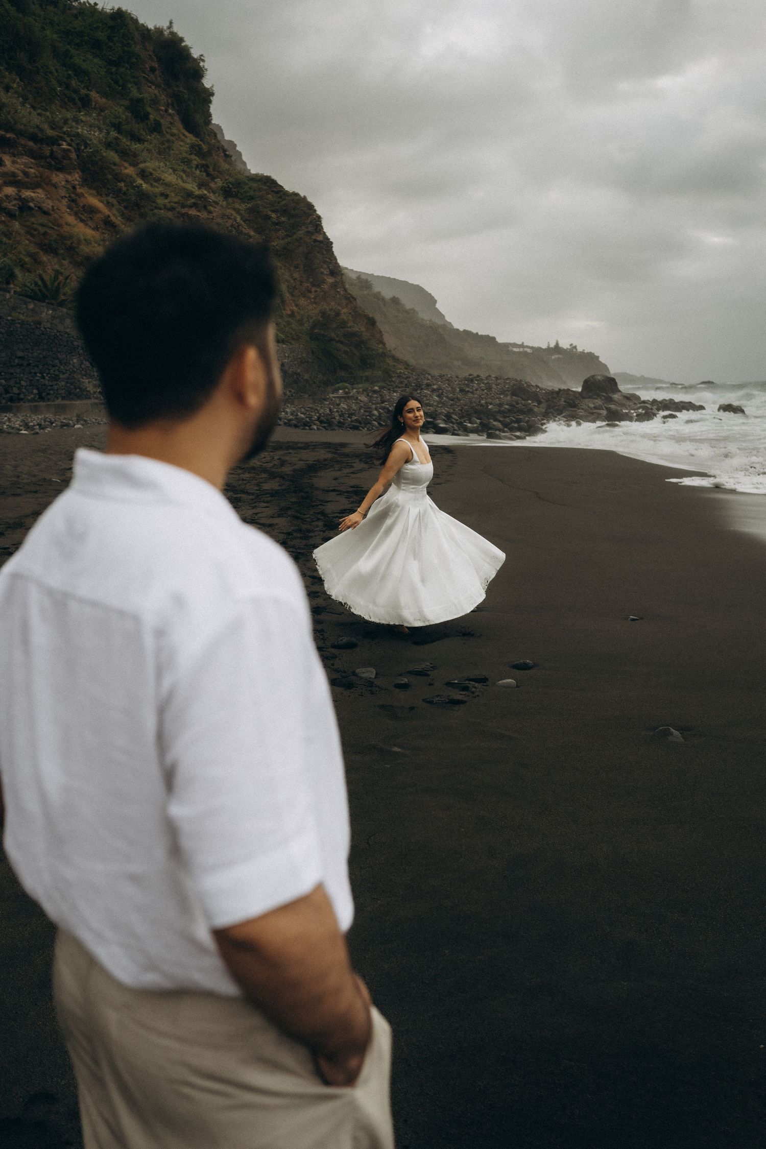 Couple on black sand beach, woman in white dress twirling near dramatic cliffs and stormy ocean waves.