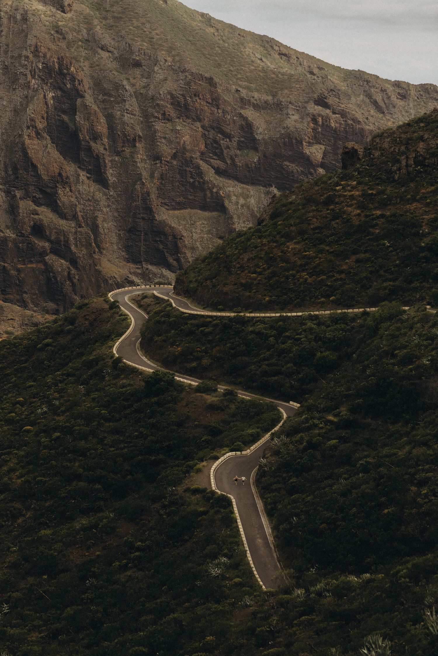 Aerial view of a winding mountain road with sharp curves cutting through lush green hillsides and rocky terrain.