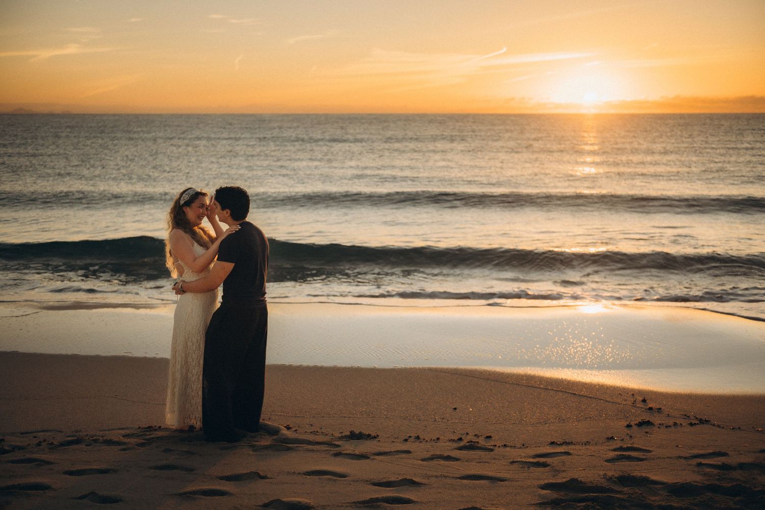 Couple kissing on sandy beach at golden sunset with ocean waves in background, romantic engagement photo.