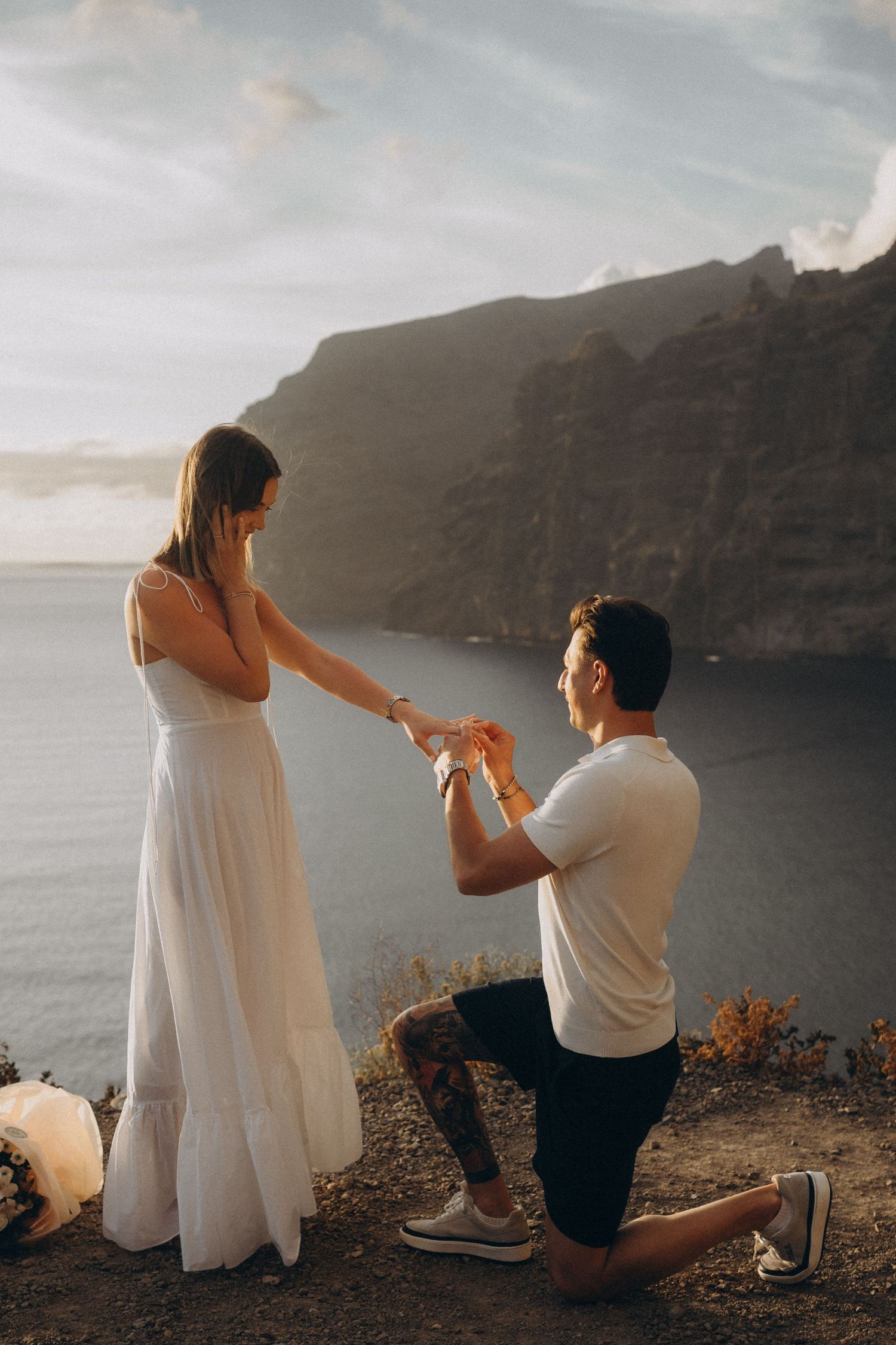 Man proposes to woman on cliff overlooking ocean at sunset, she wears white dress as he kneels with ring.