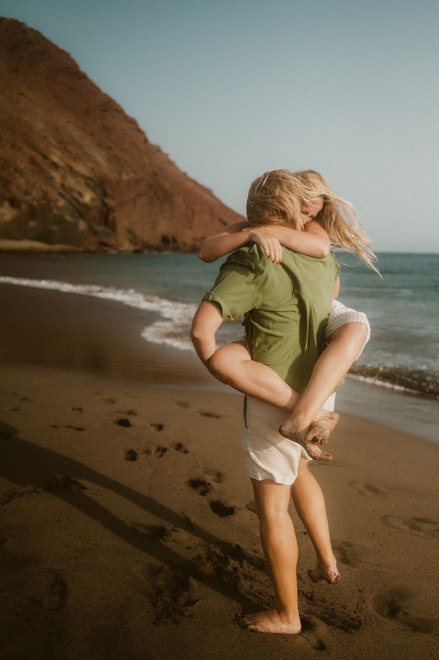 Young couple on Playa Tejita, he holds her in his arms in an embrace, ocean and Montaña Roja in the background, Tenerife couples photoshoot