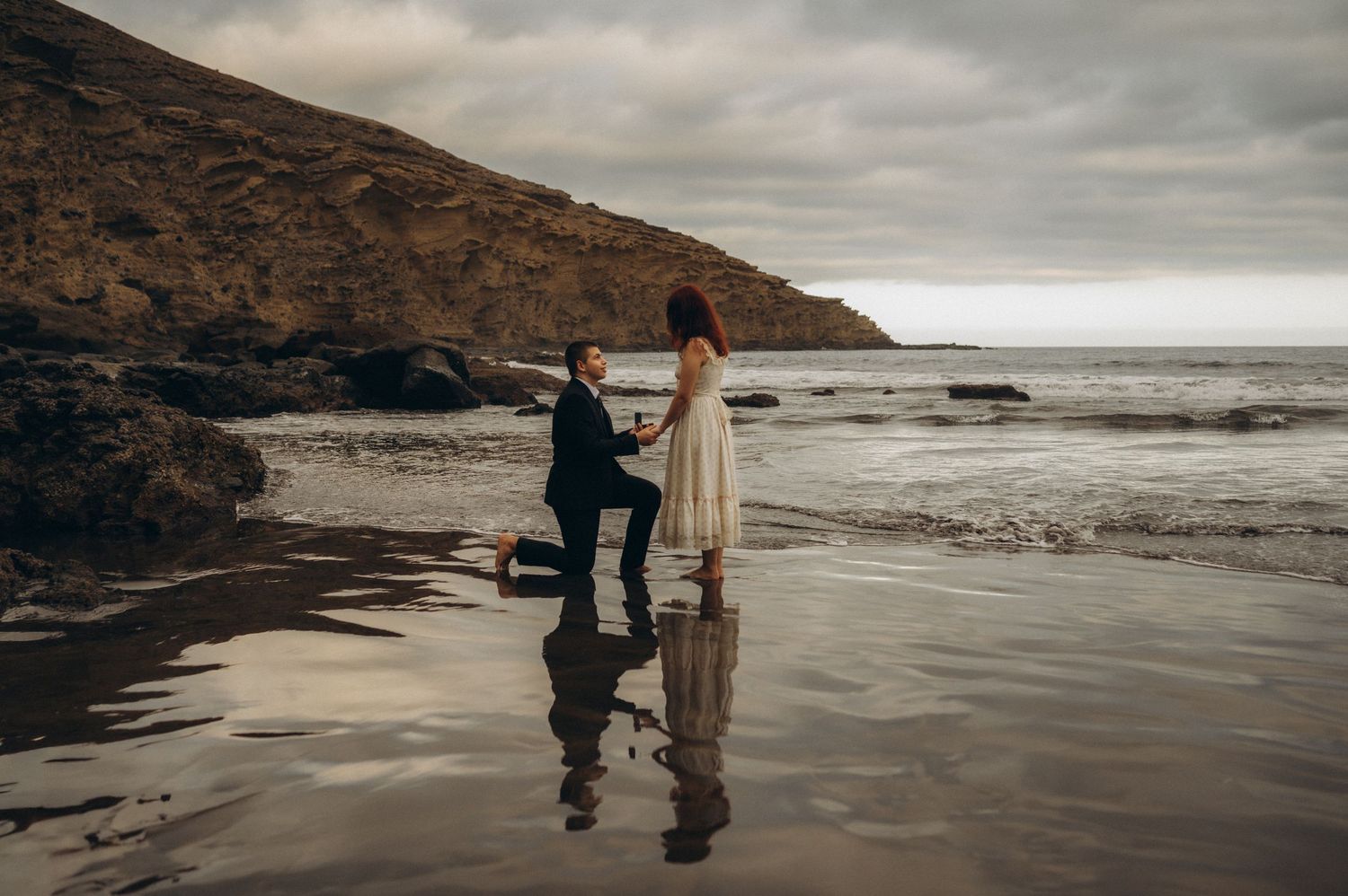 Surprise proposal in Tenerife at Playa Montaña Pelada on a reflective beach with dramatic coastal cliffs and ocean waves.
