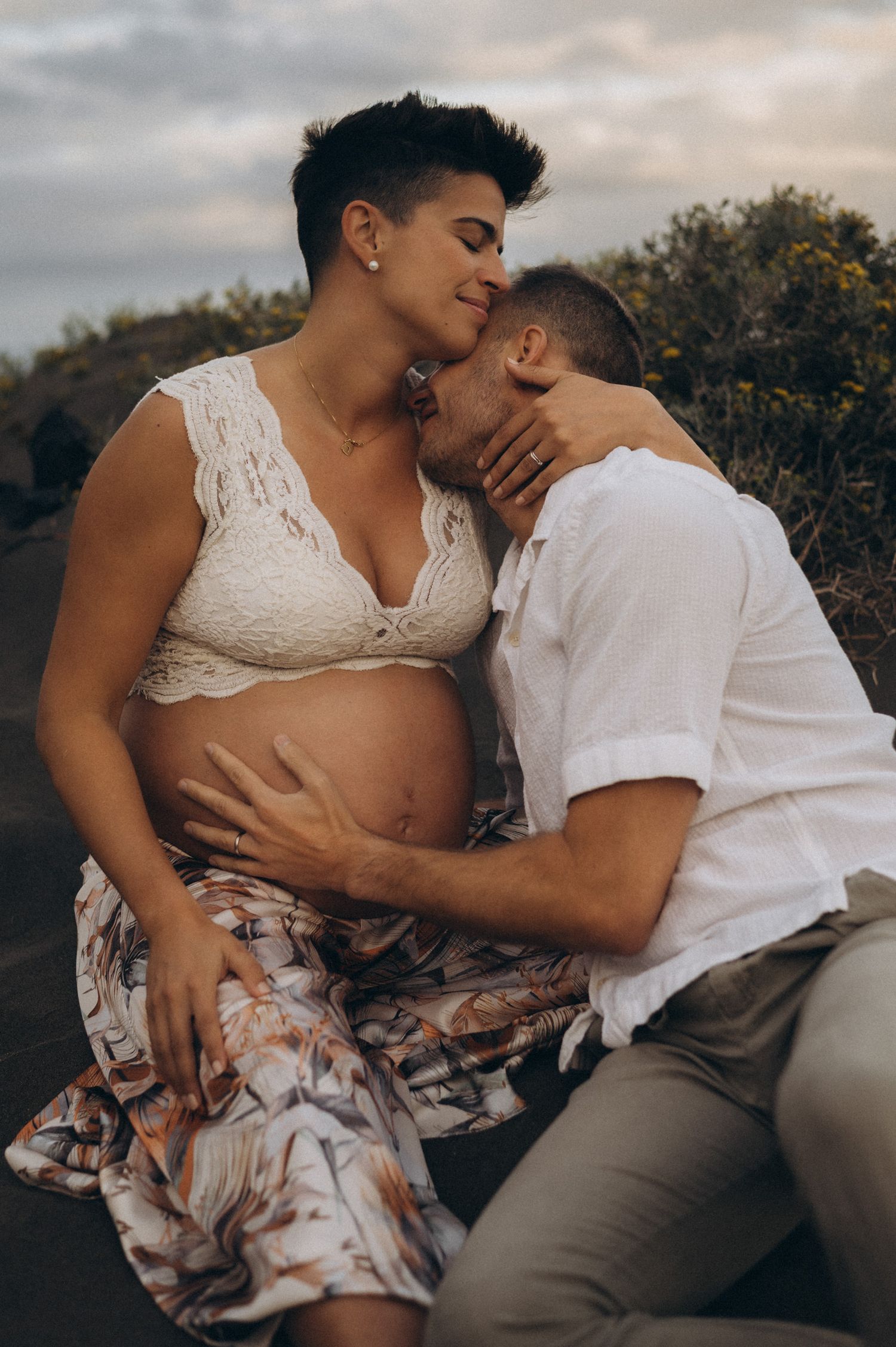 Pregnant woman in lace bralette sits outdoors with partner kissing her cheek, both enjoying a tender maternity moment.