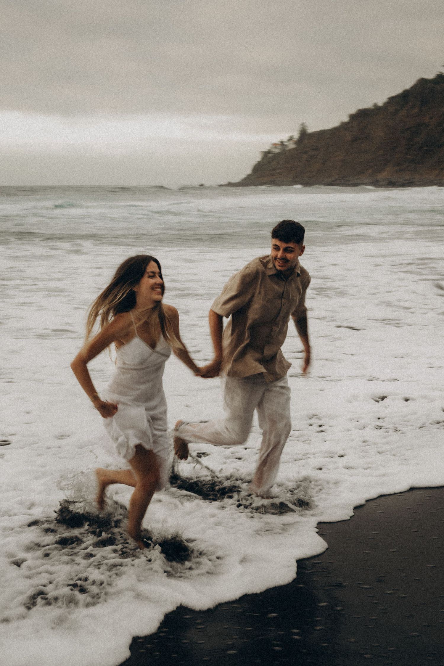 A joyful couple runs through ocean waves on a black sand beach, laughing together on a cloudy day.