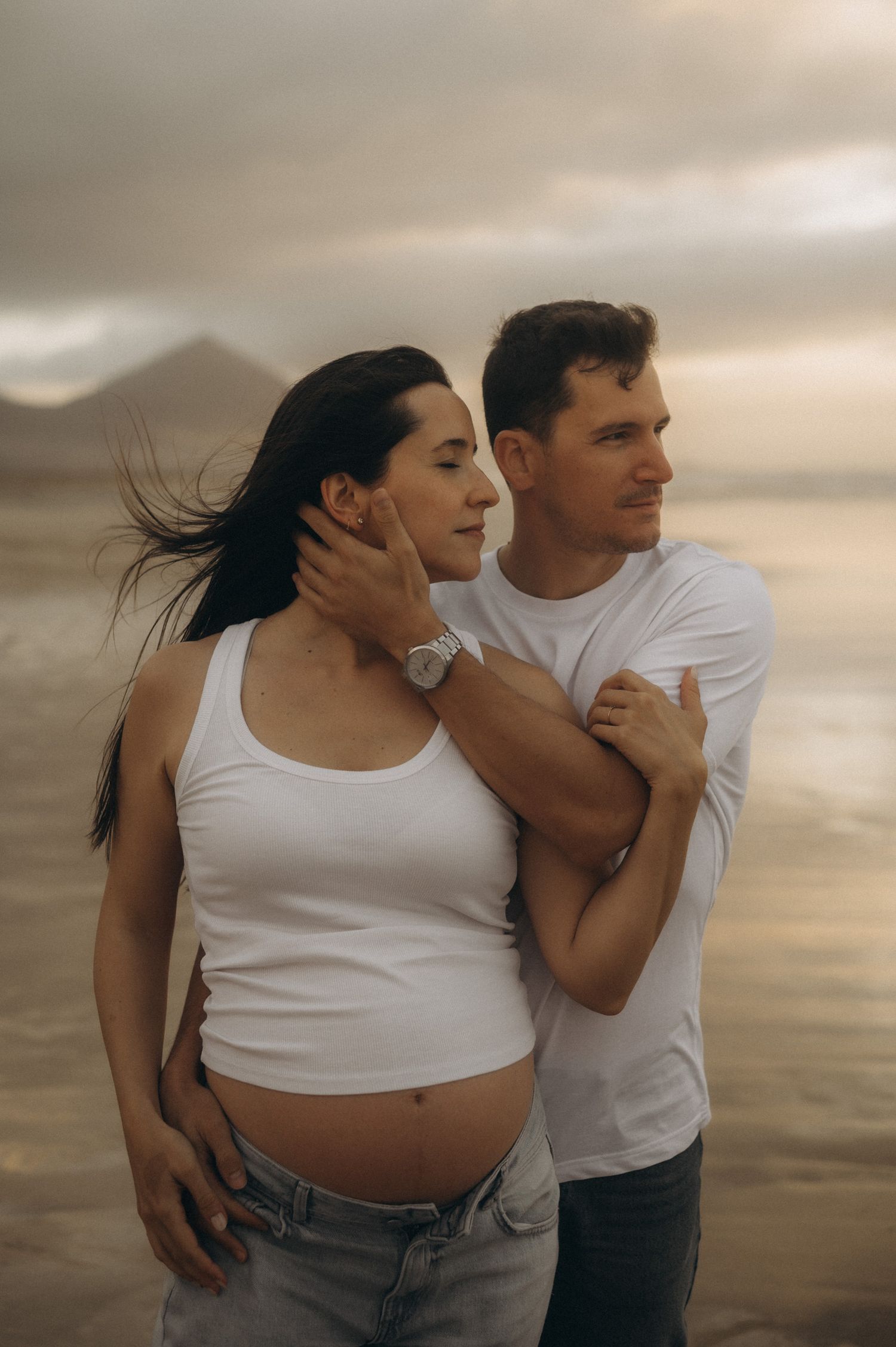 Pregnant couple embracing on a beach at sunset, wearing white tops, with mountains and dramatic sky in the background.