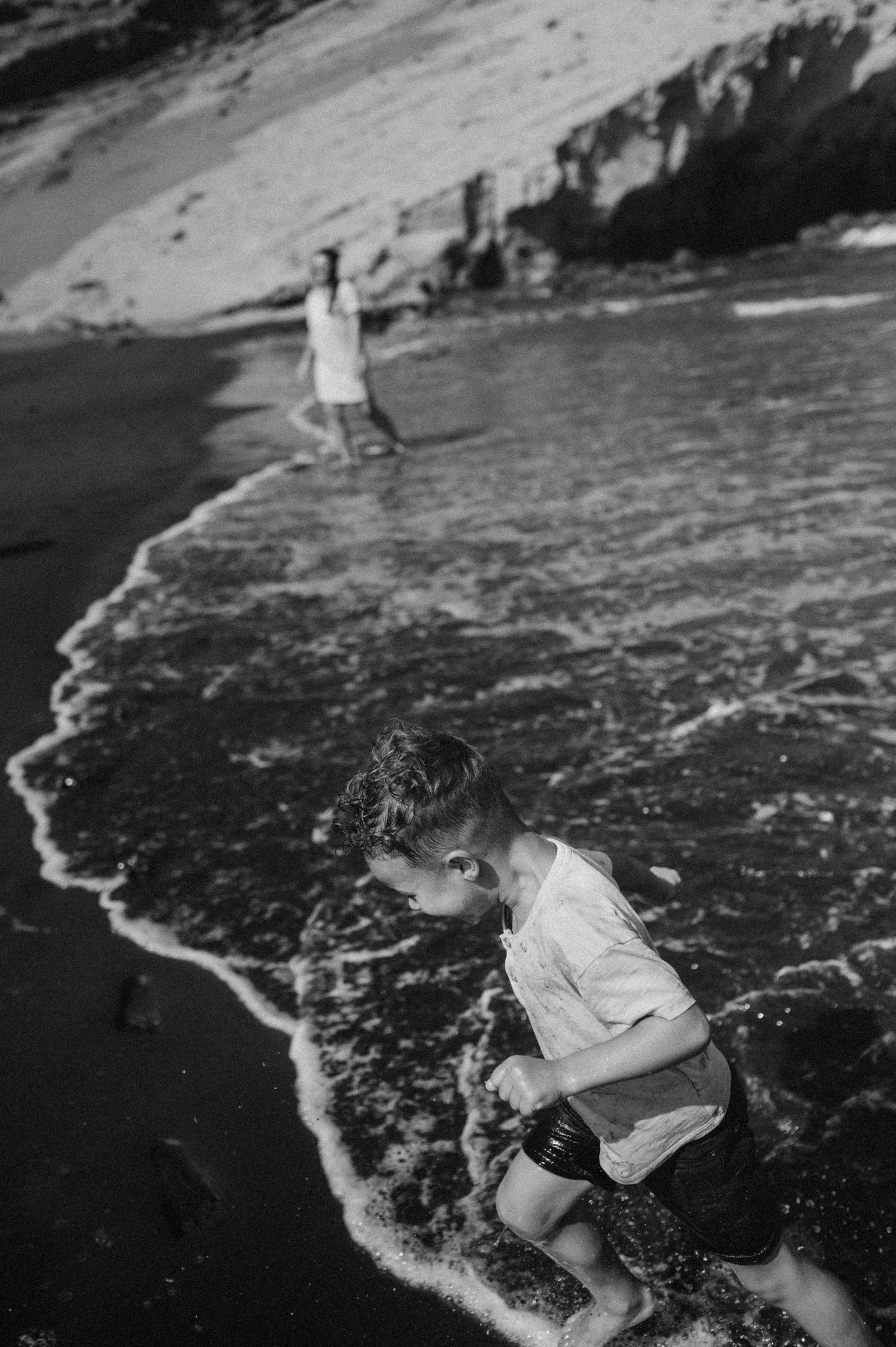 Black and white photo of mom and son playing in ocean waves along scenic Tenerife beach shoreline during family photoshoot.