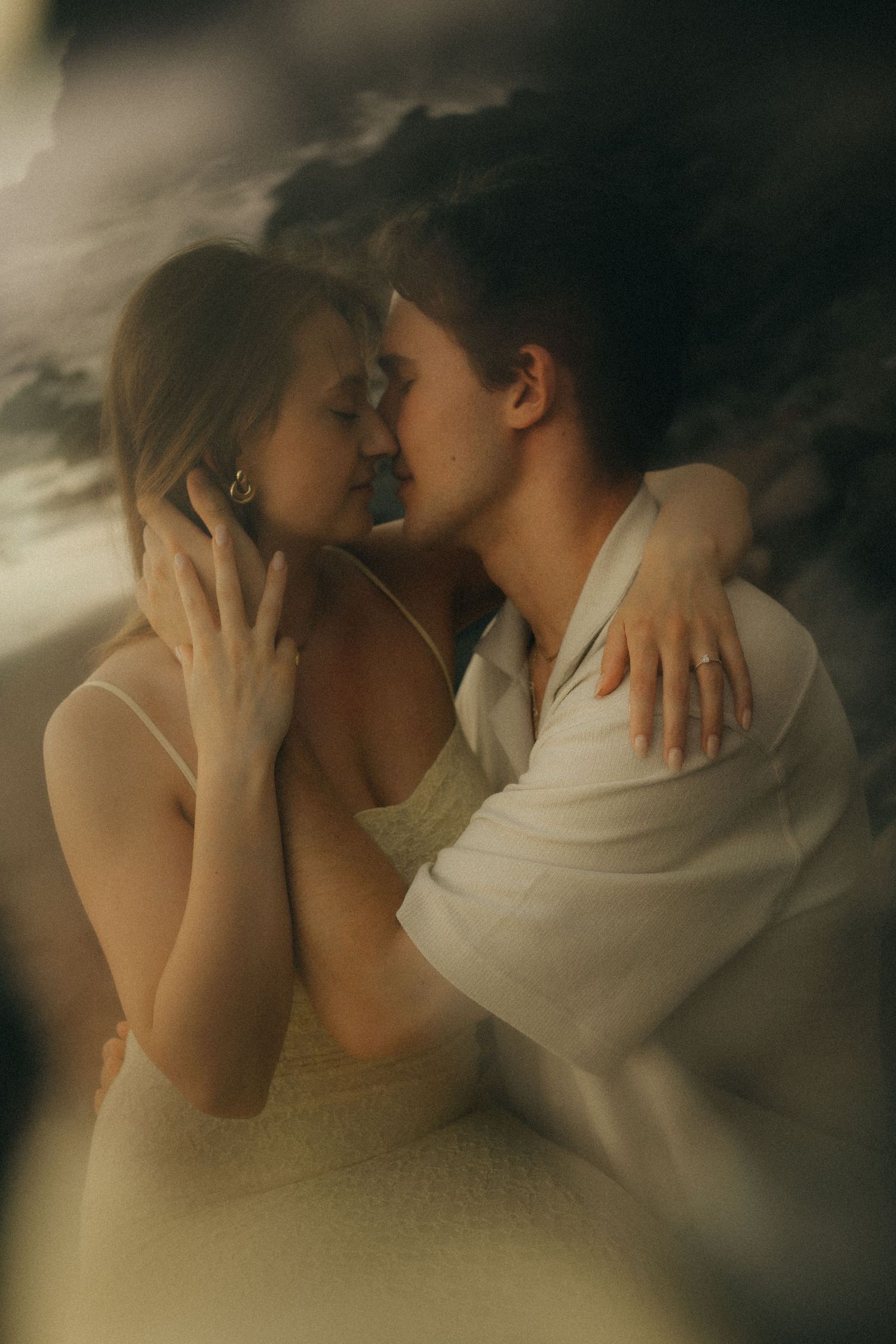 A romantic couple in white attire shares an intimate embrace against a dramatic stormy sky background.
