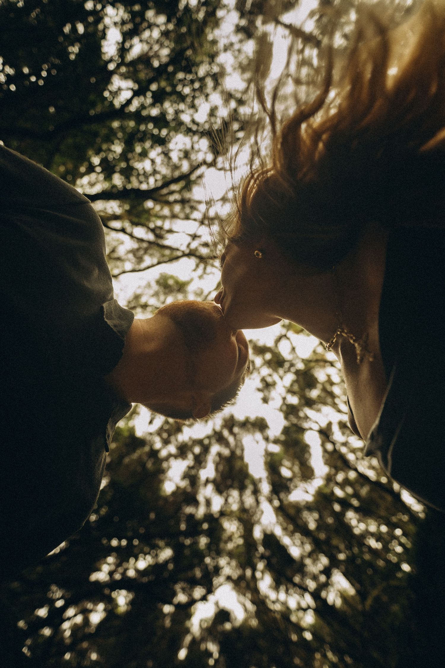 Couple sharing a romantic kiss outdoors, photographed from below with sunlit trees in the background.