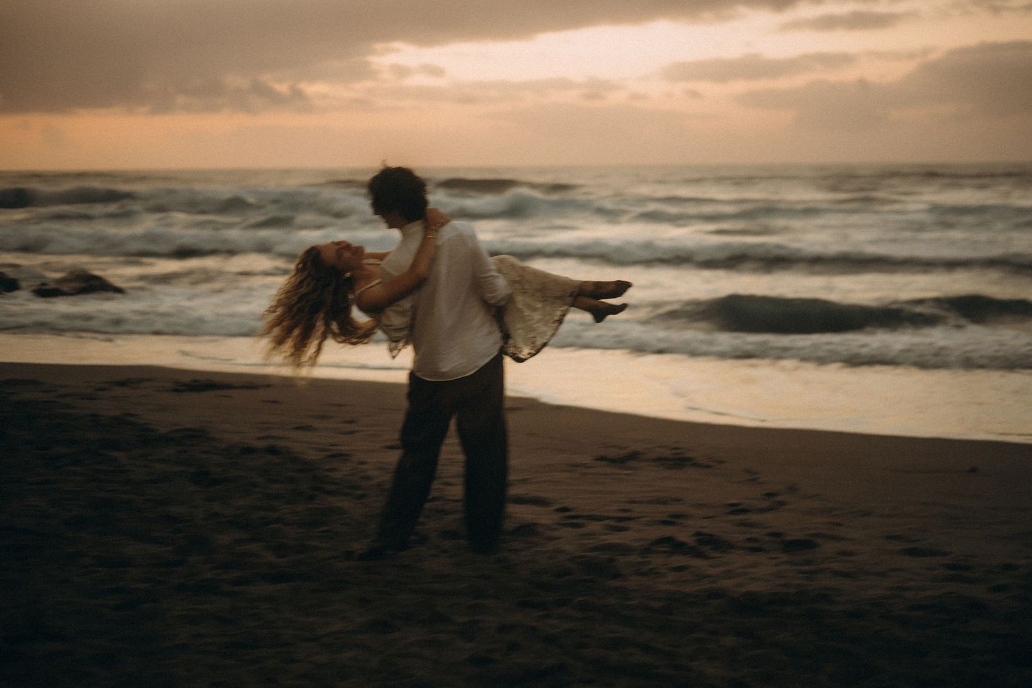 Couple dancing romantically on a sandy beach at sunset with ocean waves in the background.