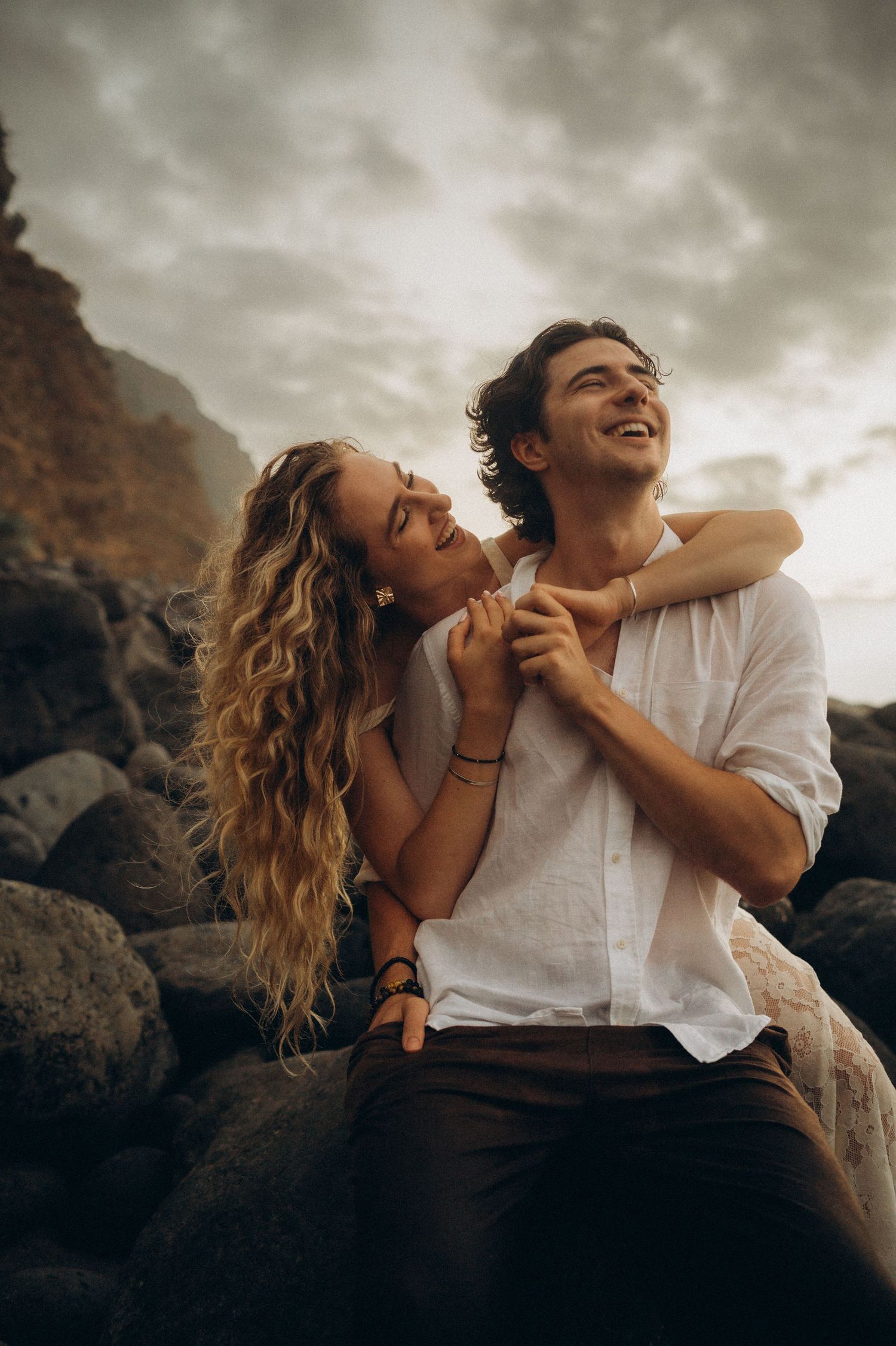 A smiling couple sitting on rocky shore, woman with long blonde hair embracing man in white shirt under cloudy sky.