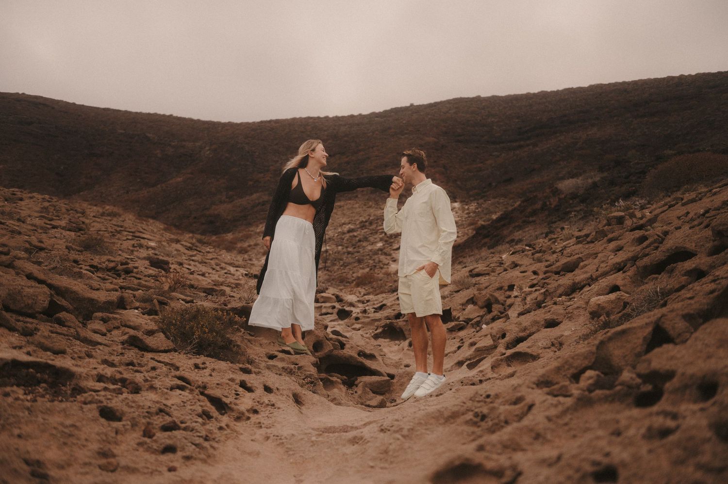 „Loving couple standing face-to-face on the rocks under Montaña Roja, he gently kissing her hand during a romantic moment, Tenerife