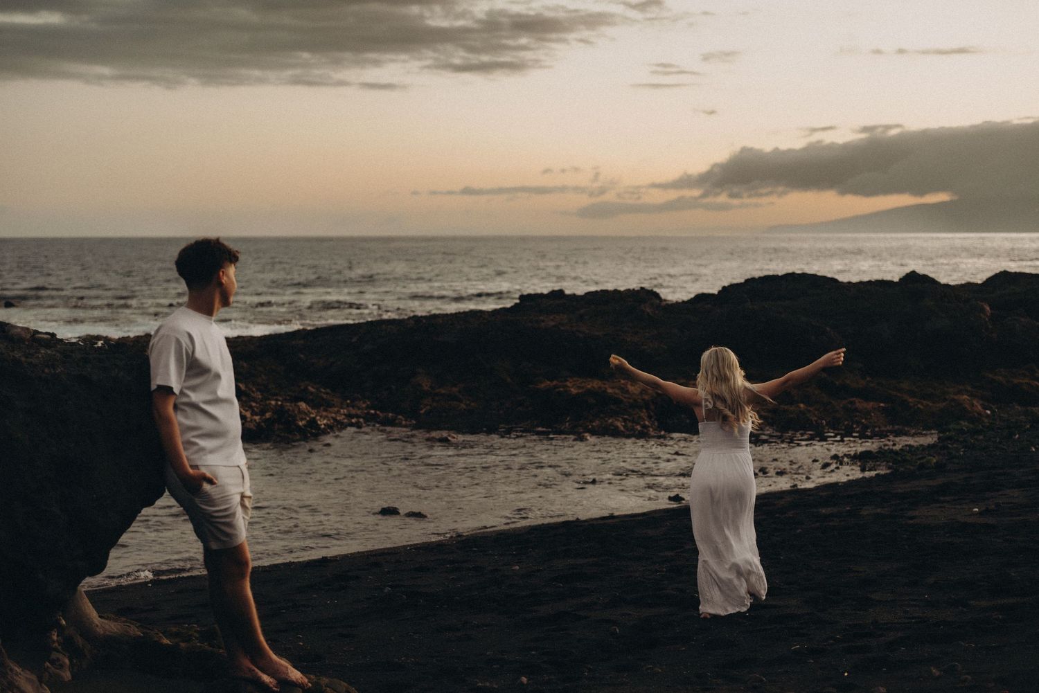 Couple standing on rocky beach at sunset, woman in white dress with arms outstretched near ocean waves.