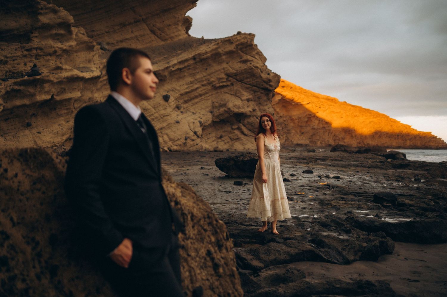 A couple in formal attire poses on a rocky beach at sunset, with golden light illuminating dramatic cliff walls behind them.