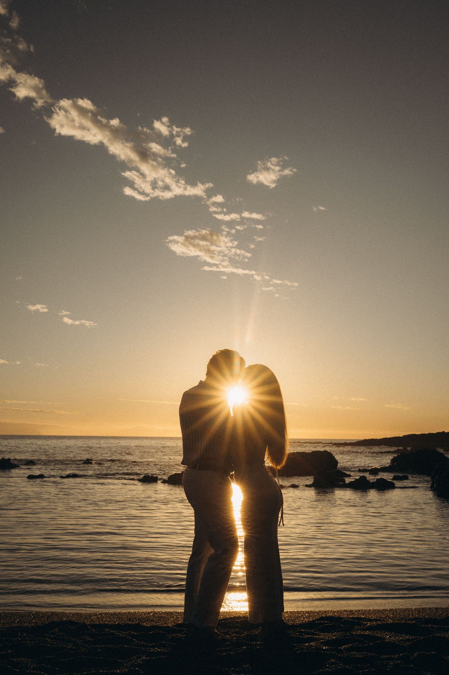 Silhouette of a couple kissing on a beach at sunset with golden sun rays shining between them.