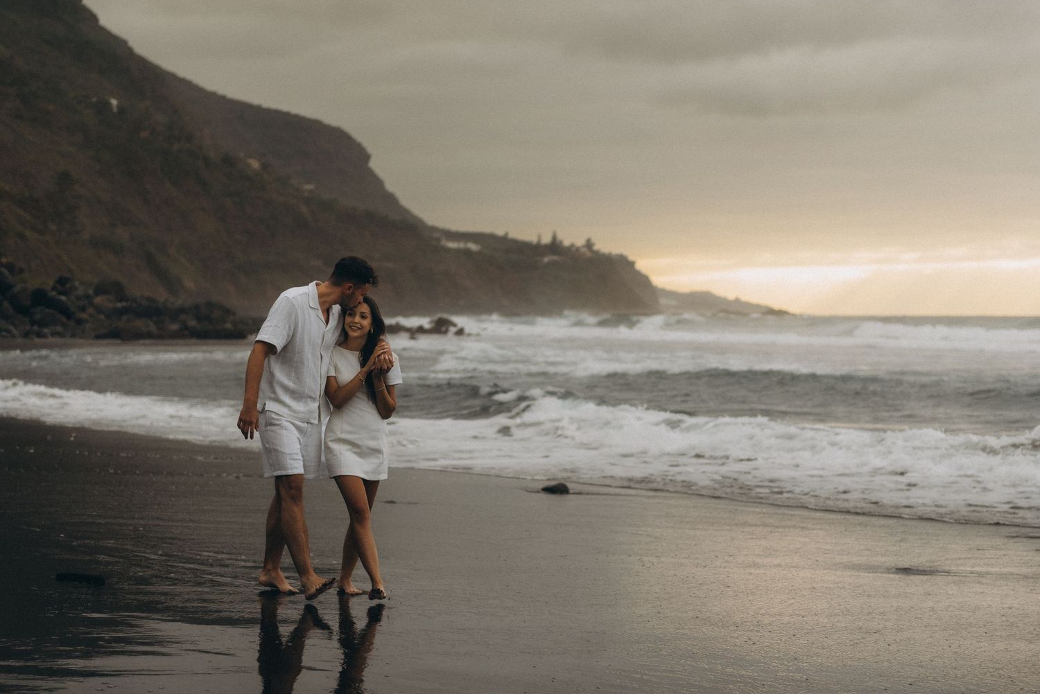 Couple walking hand in hand on a black sand beach at sunset with dramatic ocean waves and cliffs.