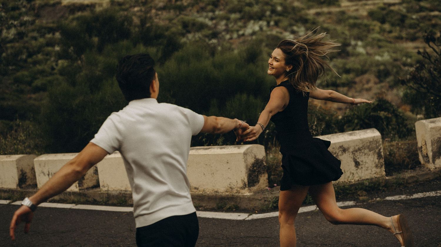 A couple joyfully runs hand-in-hand on a road surrounded by lush green hills, capturing a romantic and carefree moment.