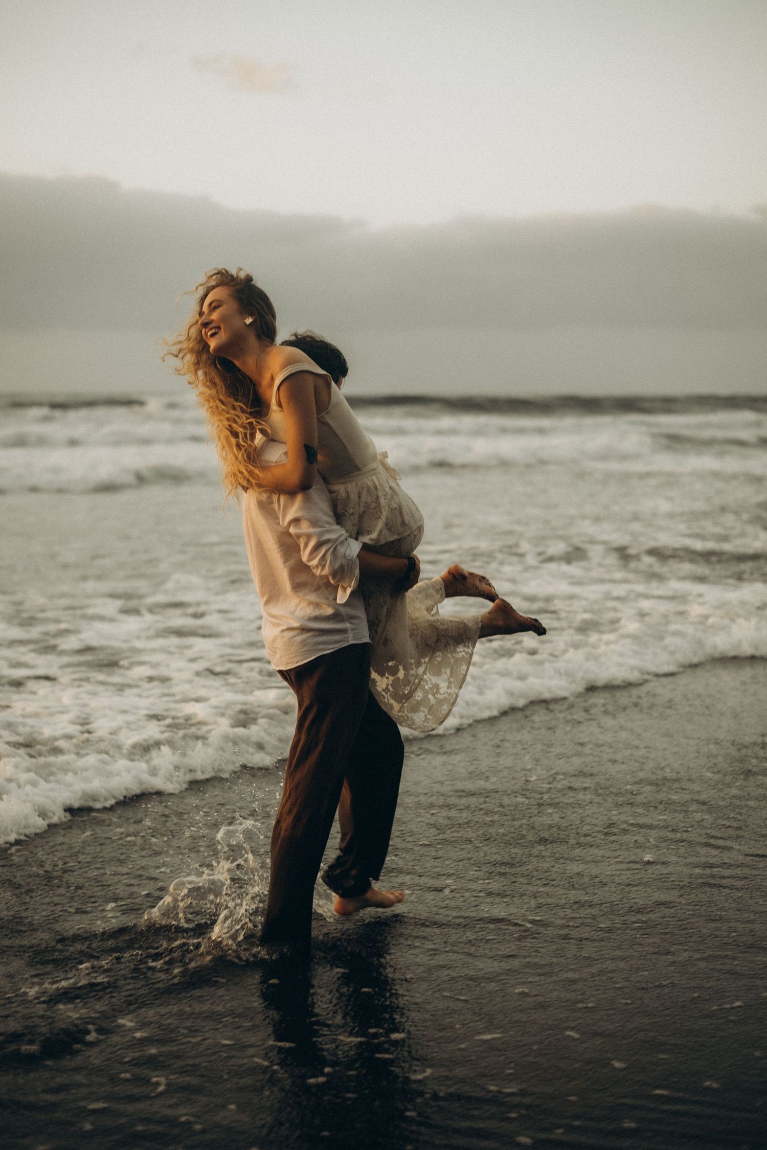 Couple embracing joyfully on a black sand beach at sunset, waves crashing around their feet.
