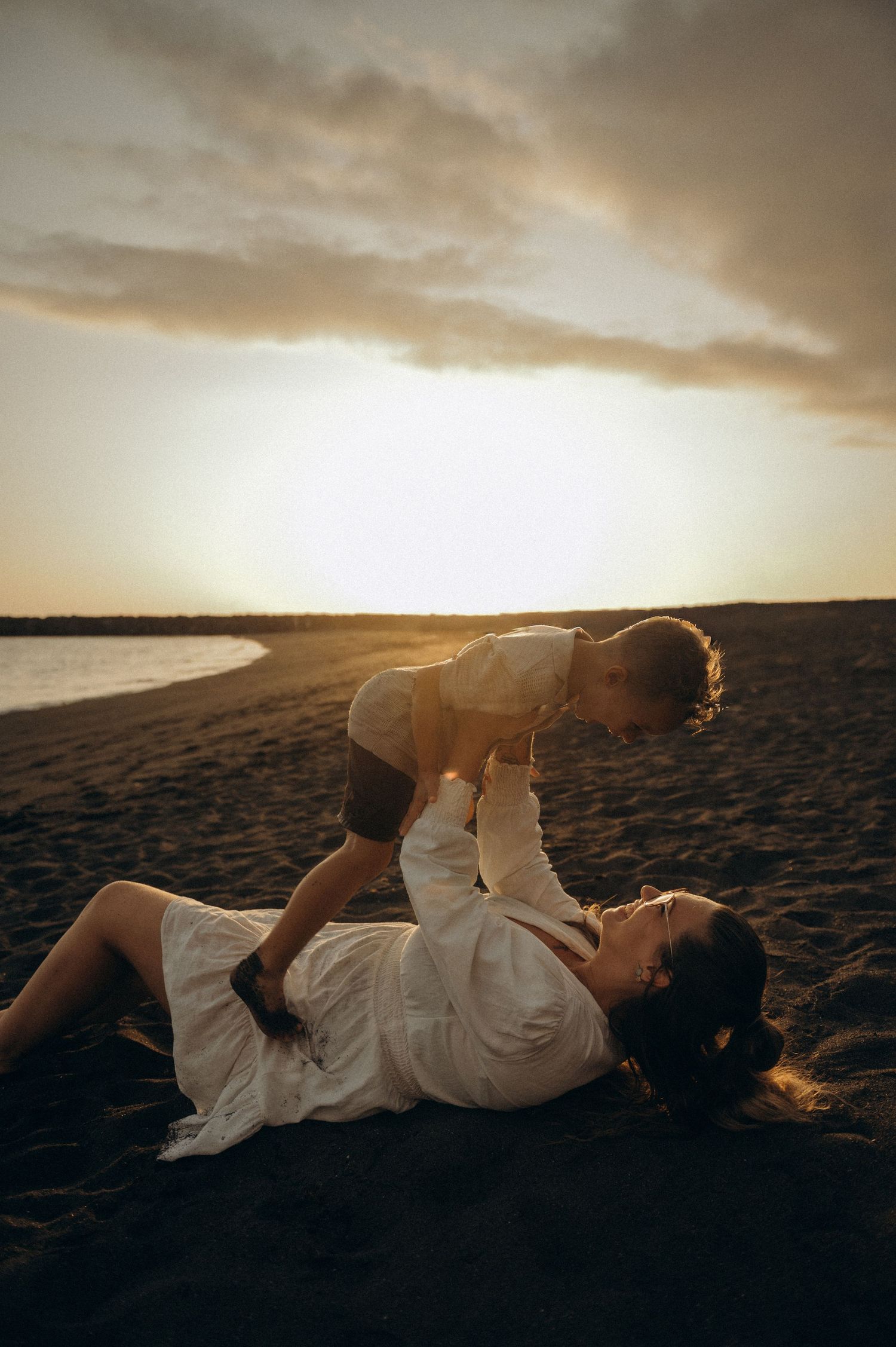 Mother lying on black sand lifting toddler in arms, both laughing at sunset, Playa del Duque