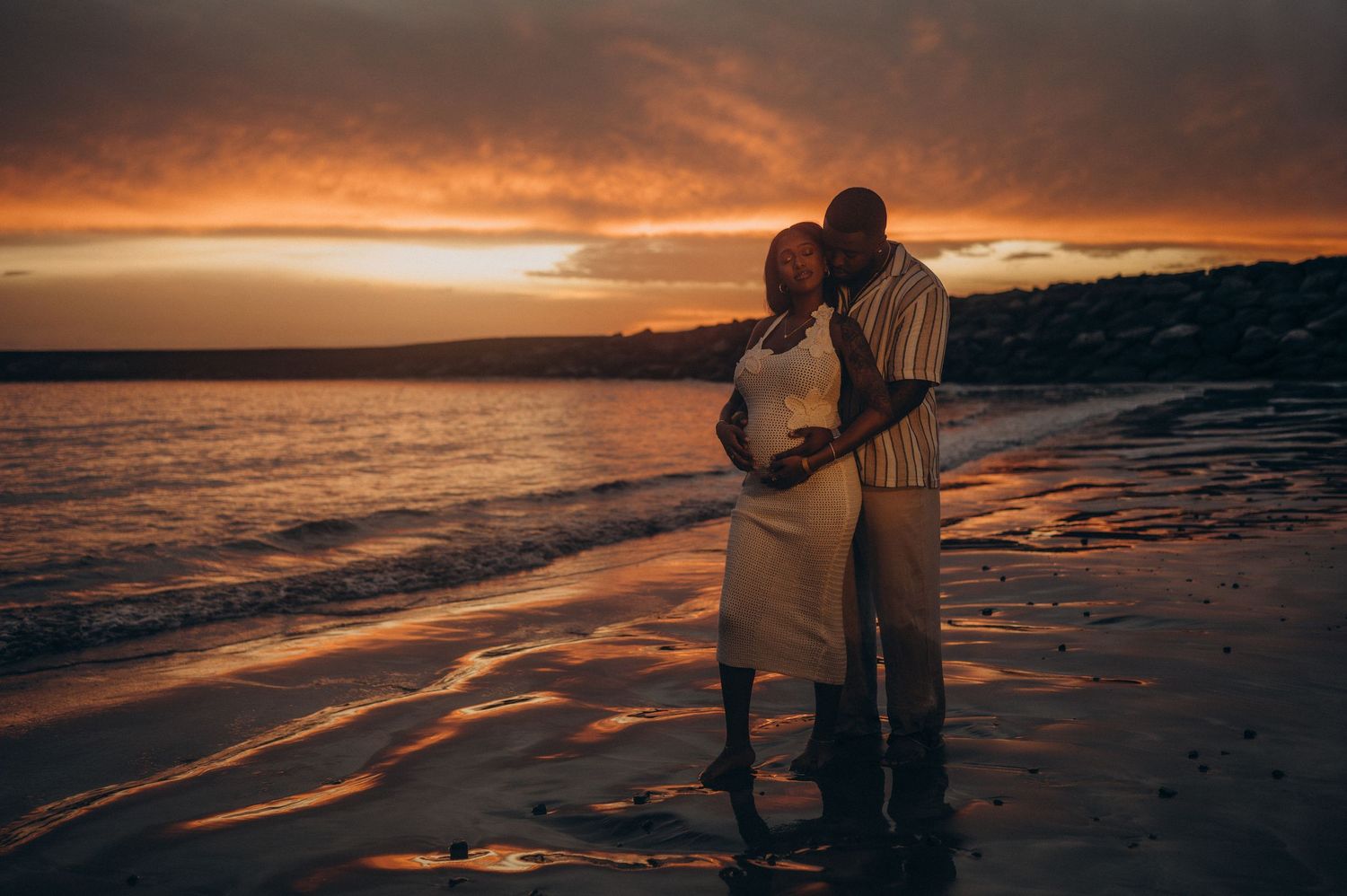Husband hugging wife from behind holding her pregnant belly at sunset, Playa Fañabé