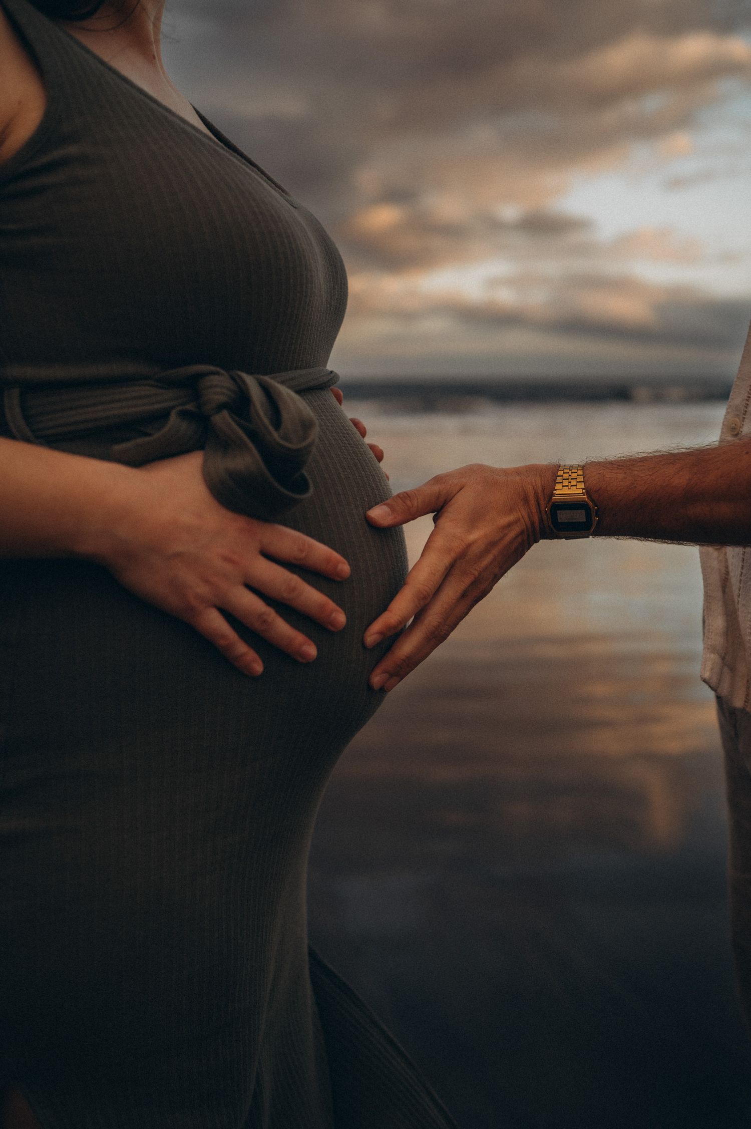 Pregnant woman in grey dress with partner's hand on baby bump at sunset beach.