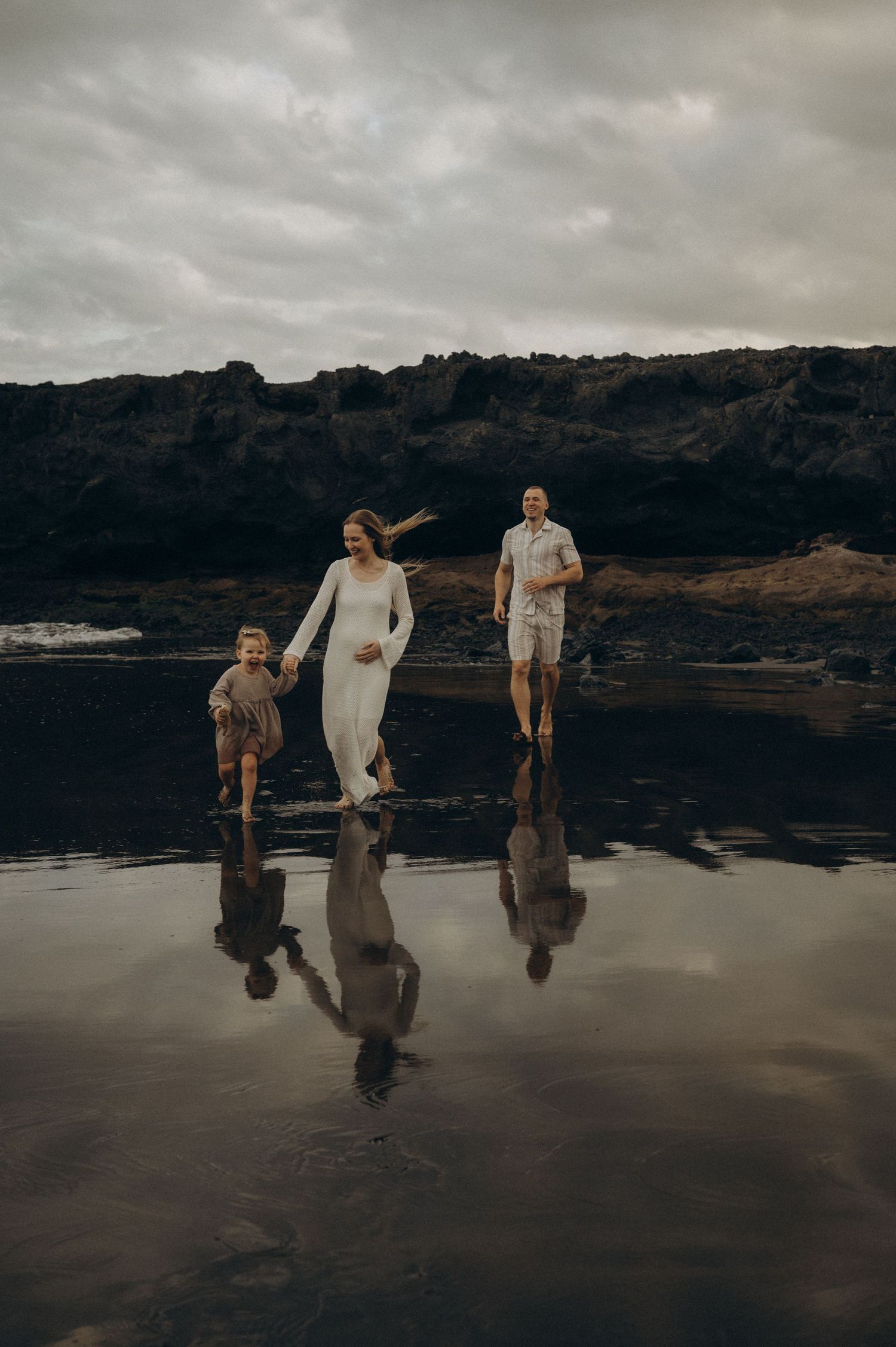 Family of three walking on reflective black sand beach with dramatic rocky coastline and cloudy sky.