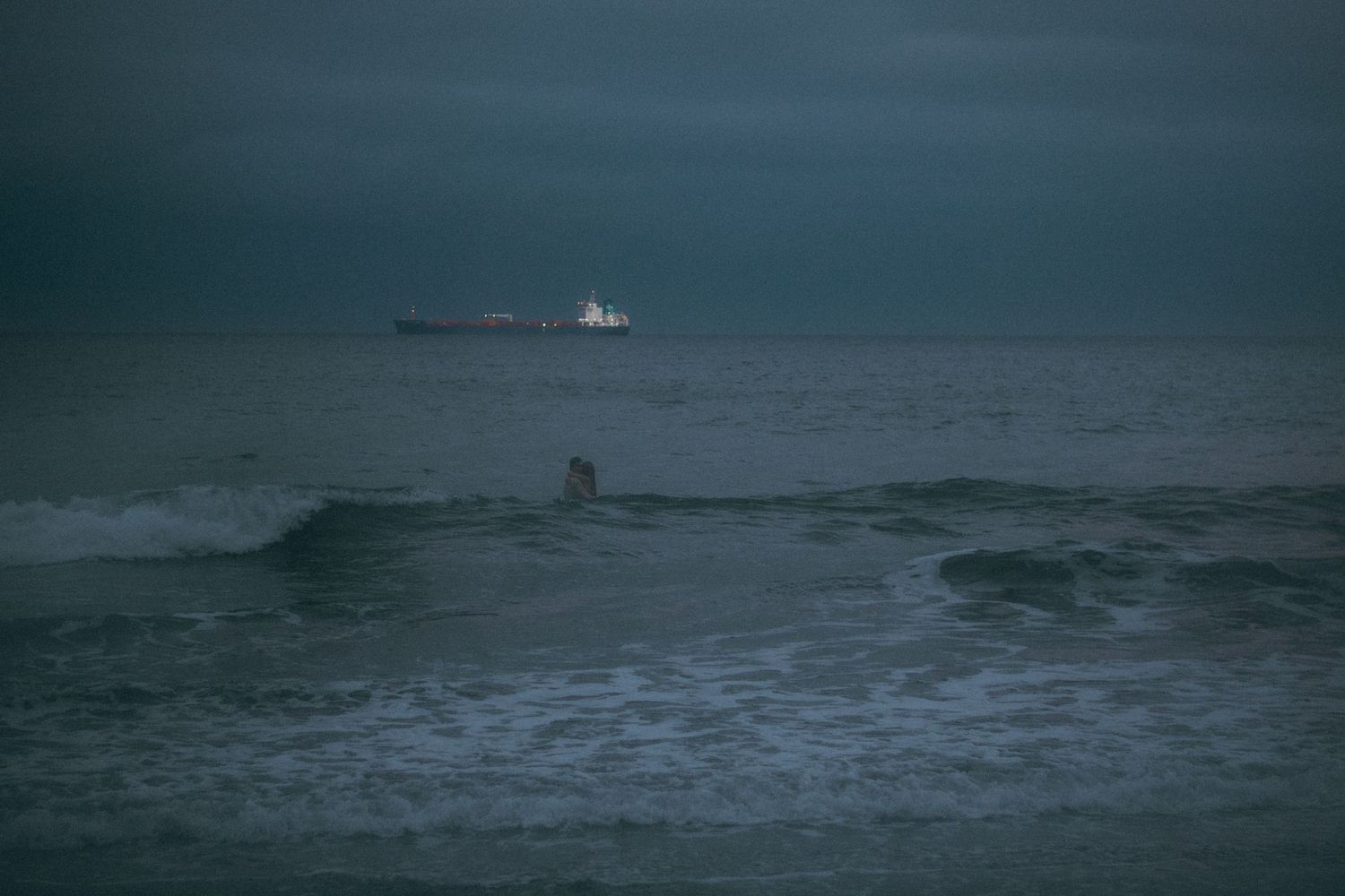Surfer riding waves in a dark stormy ocean with a large cargo ship visible on the horizon under overcast skies.