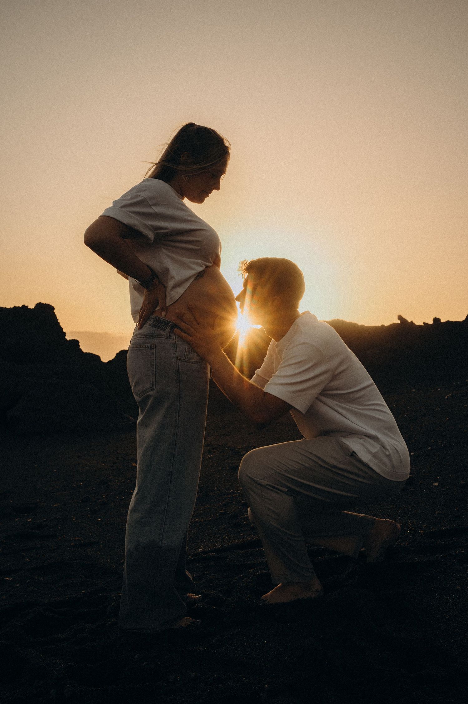 Couple sharing a tender maternity moment at sunset on rocky terrain, sun flare glowing between them.