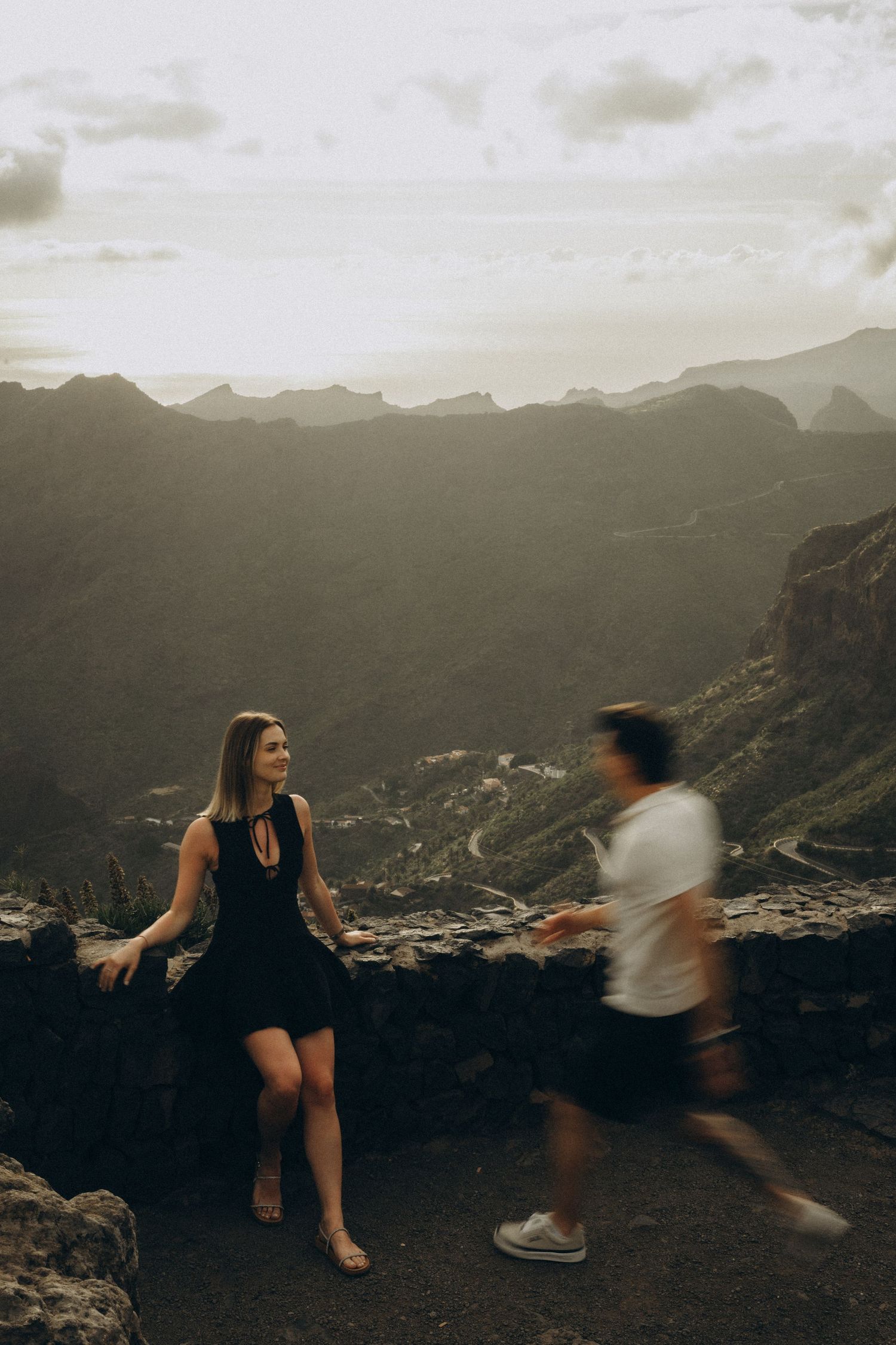 Couple at a scenic mountain viewpoint at sunset, woman in black dress leaning on stone wall with misty valleys behind.