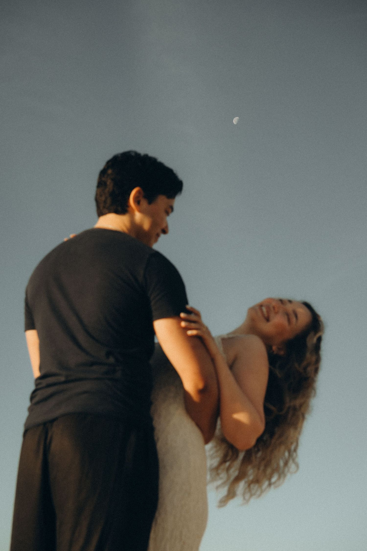 Couple dancing romantically under a crescent moon at dusk, woman laughing joyfully in a white dress.