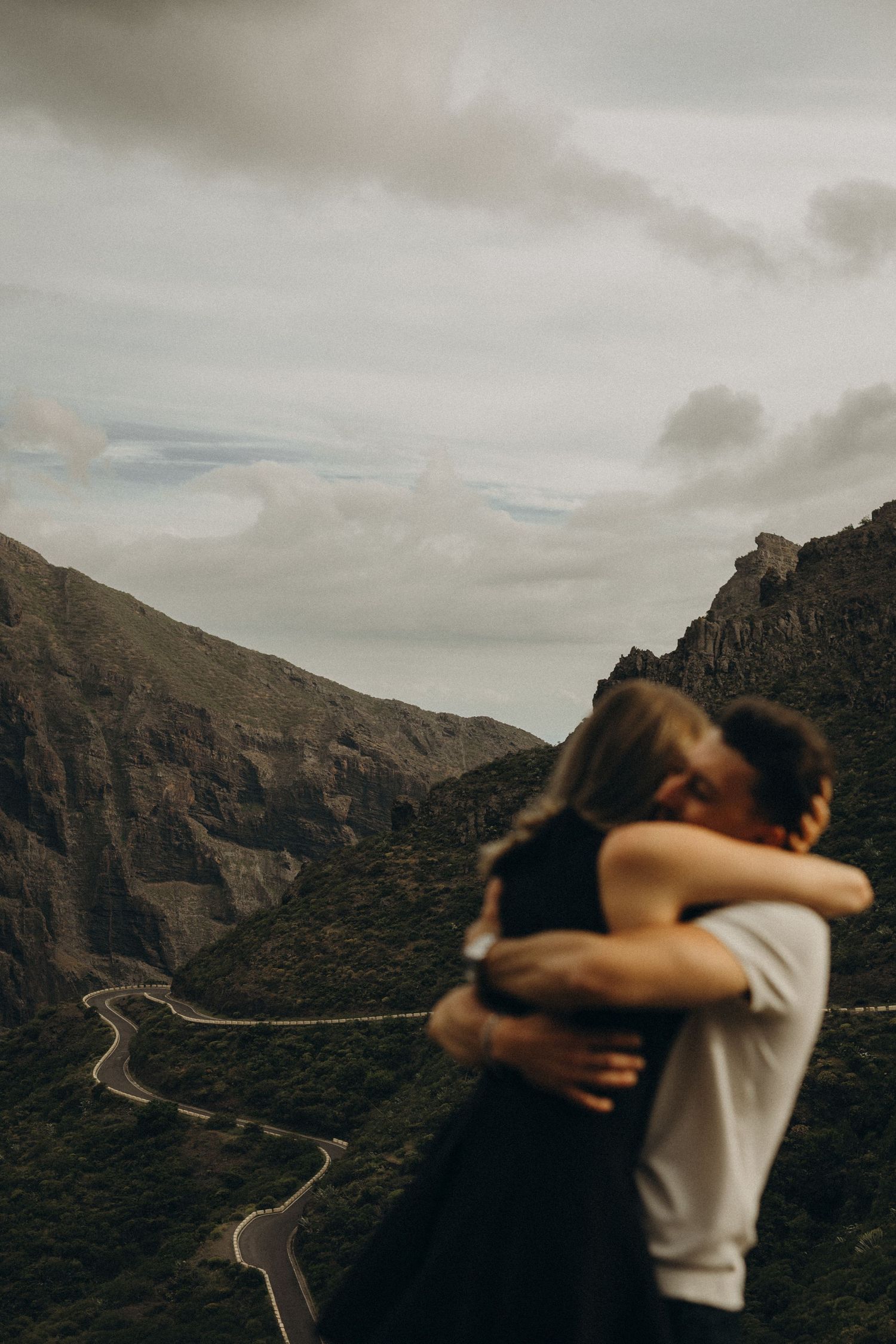 Couple embracing and kissing on a mountain overlook with a winding road and dramatic cloudy sky in the background.