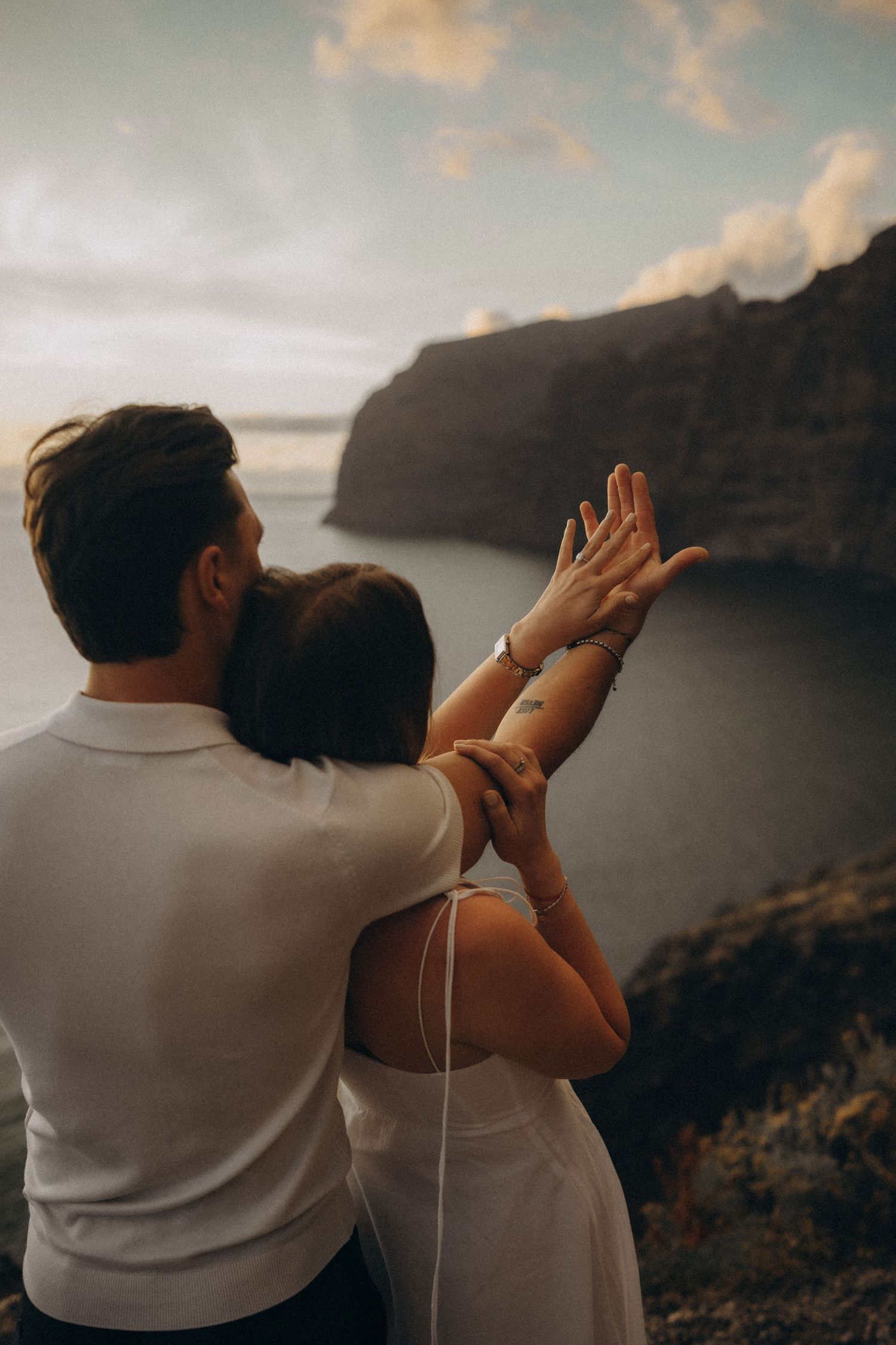 Couple embracing at sunset on Santorini cliffs, woman displaying engagement ring with ocean views.