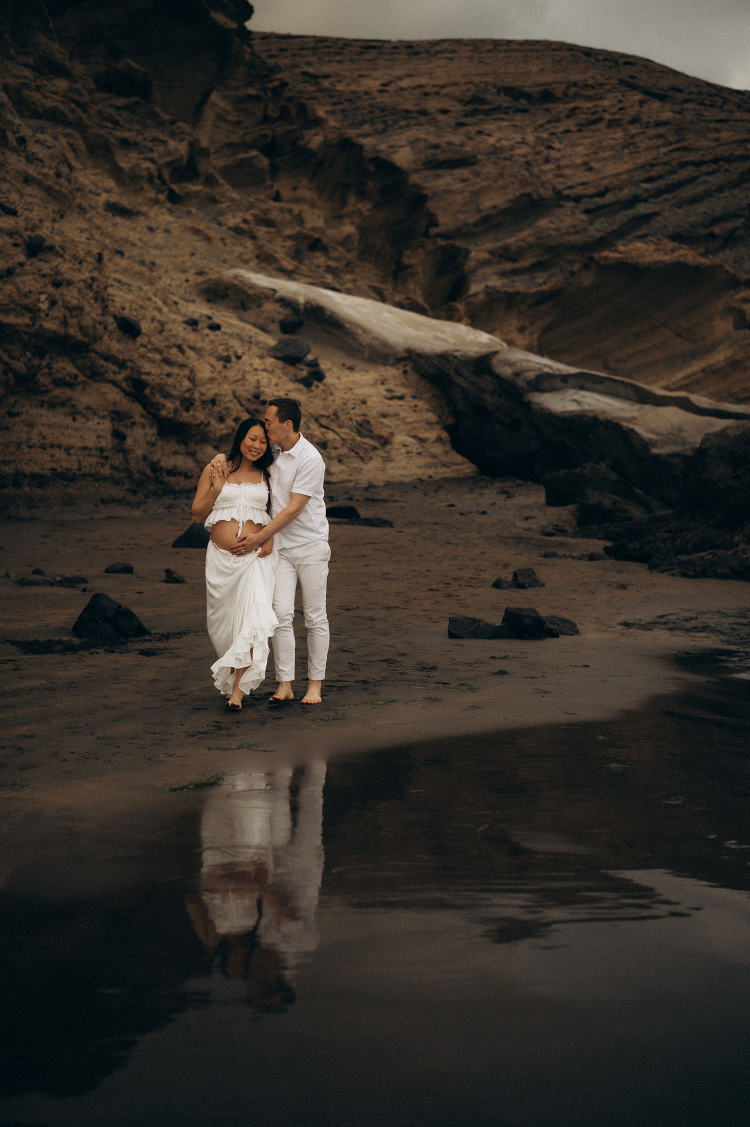 Couple in white embracing on dark beach with rocky cliffs, maternity photoshoot with stunning reflection in wet sand.