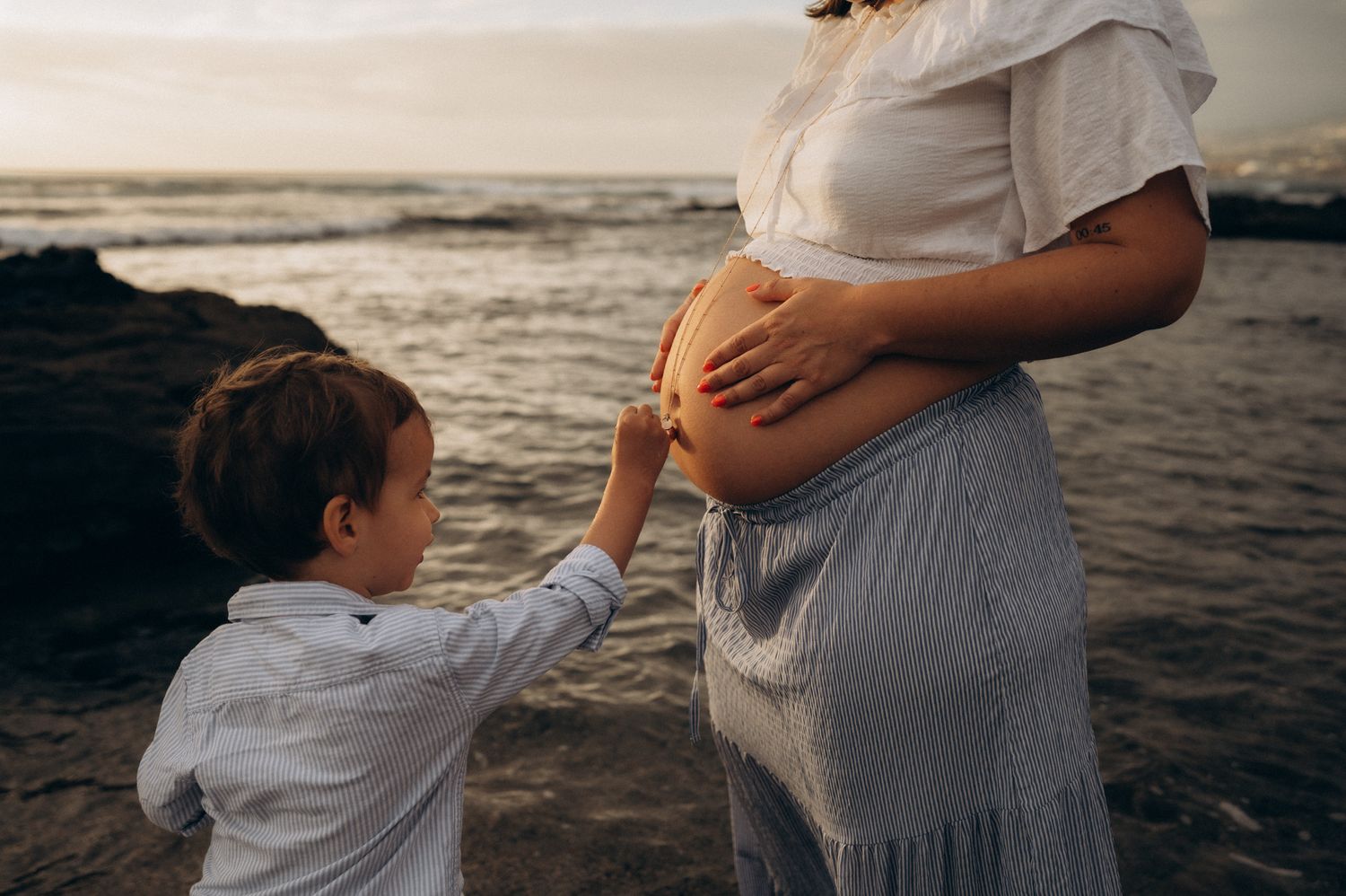 A young boy touches a pregnant woman's belly on a rocky beach at sunset.