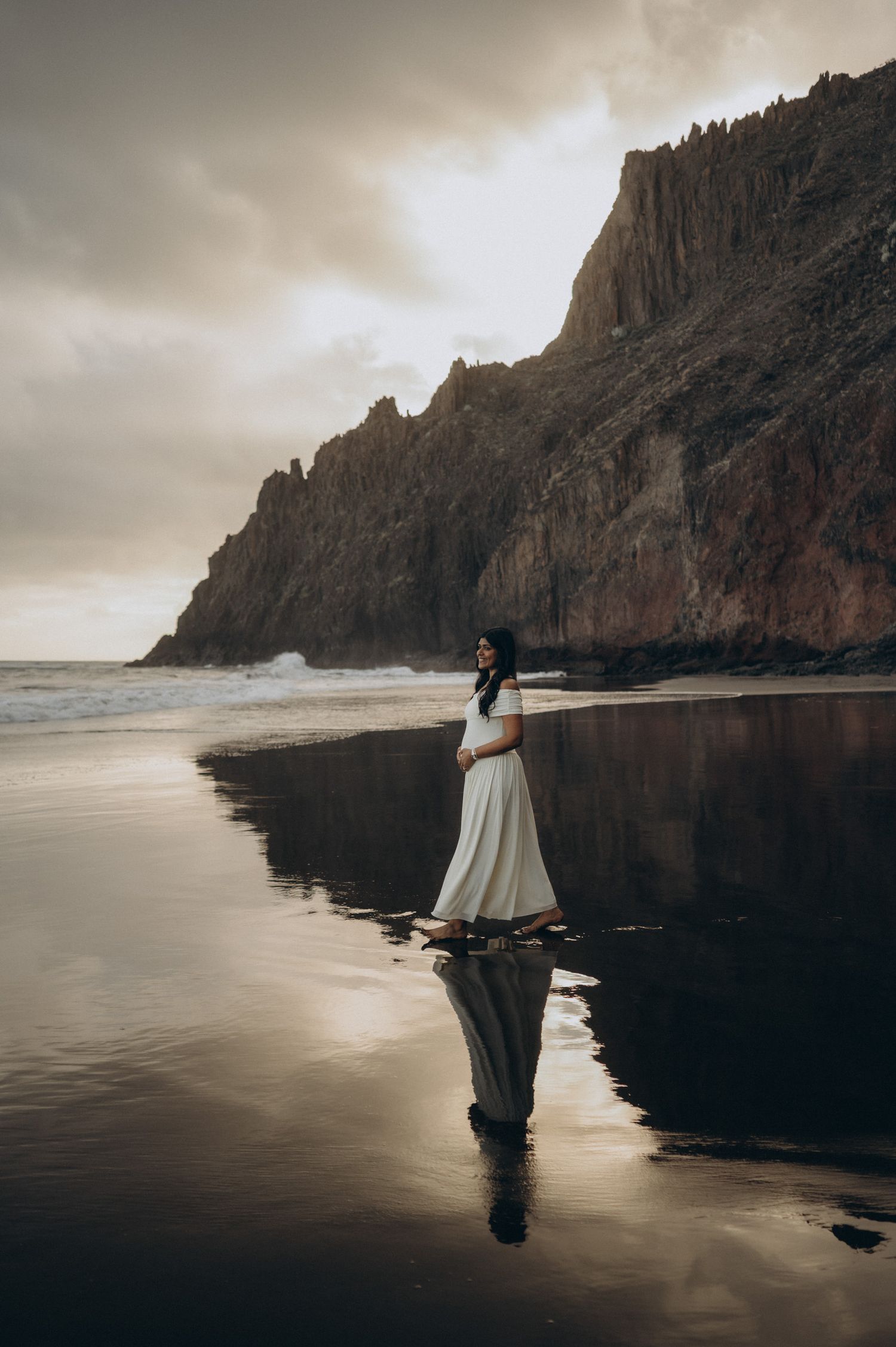 Pregnant woman in white dress stands on reflective black sand beach at dusk with dramatic cliffs behind her.