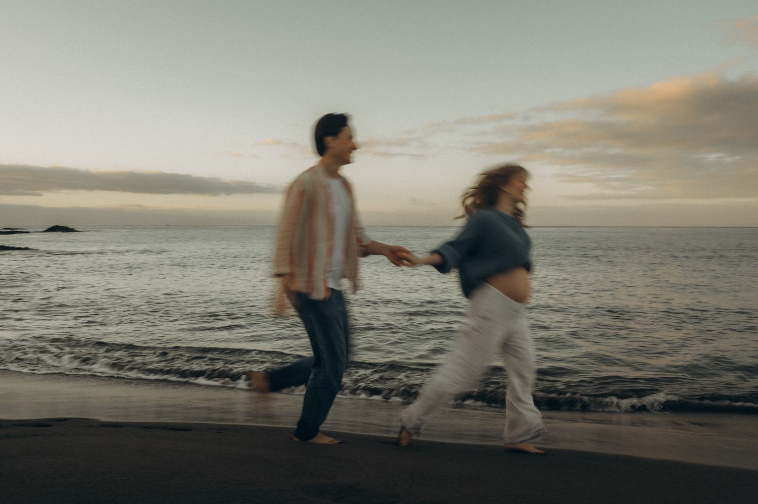 Couple holding hands running along a sandy beach at sunset with ocean waves in the background.