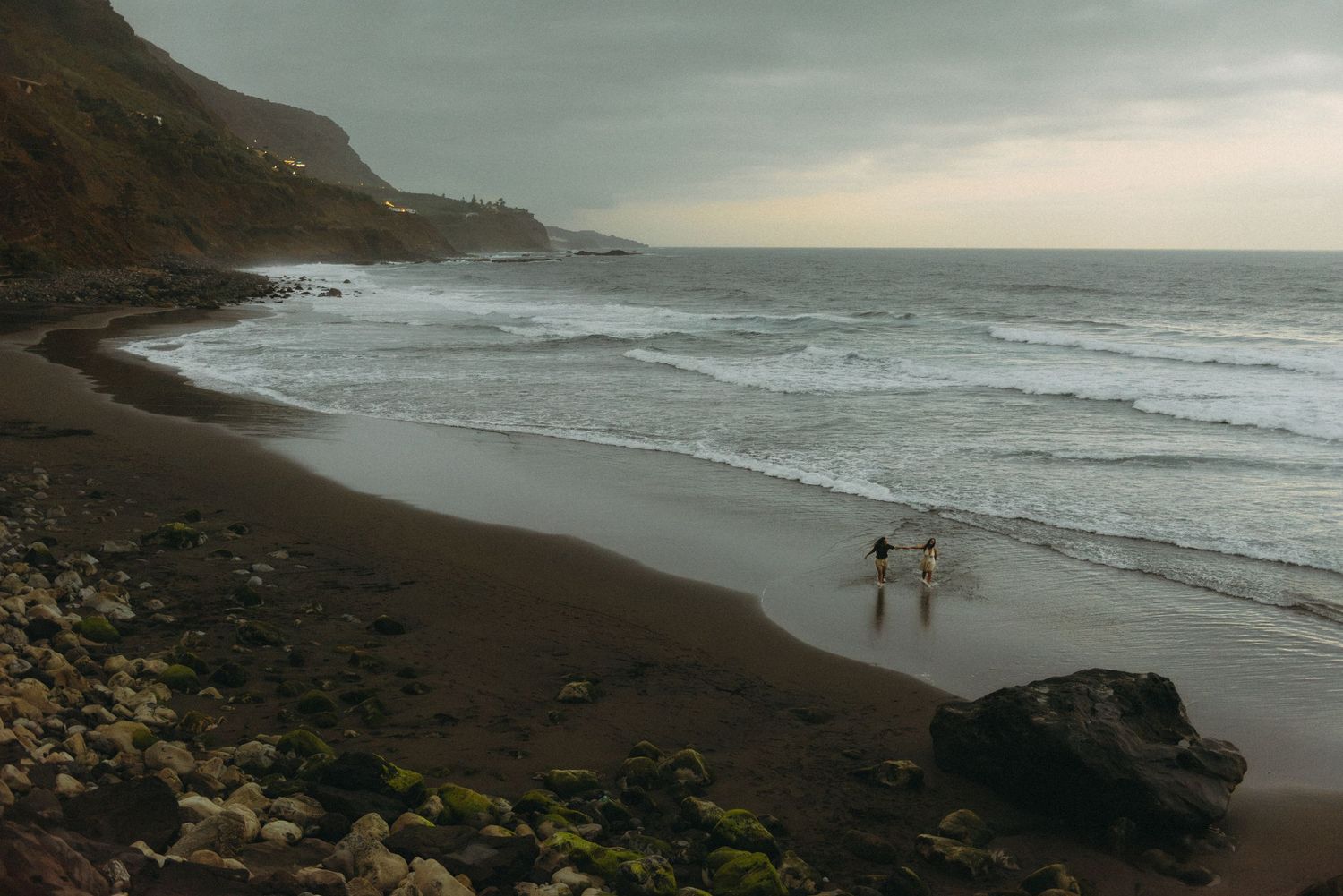 Two people walk along a dramatic dark sand beach with rocky cliffs and moody ocean waves at dusk.