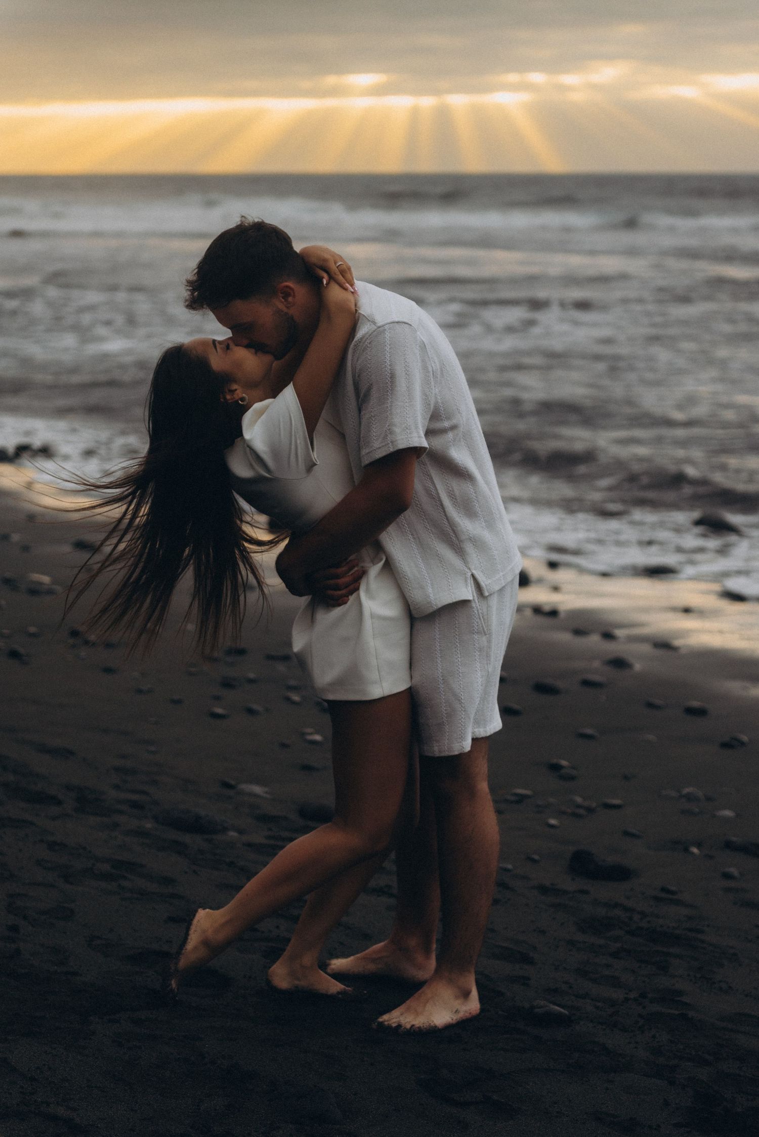 Couple kissing on black sand beach at sunset, wearing white linen outfits, with dramatic sun rays over ocean.