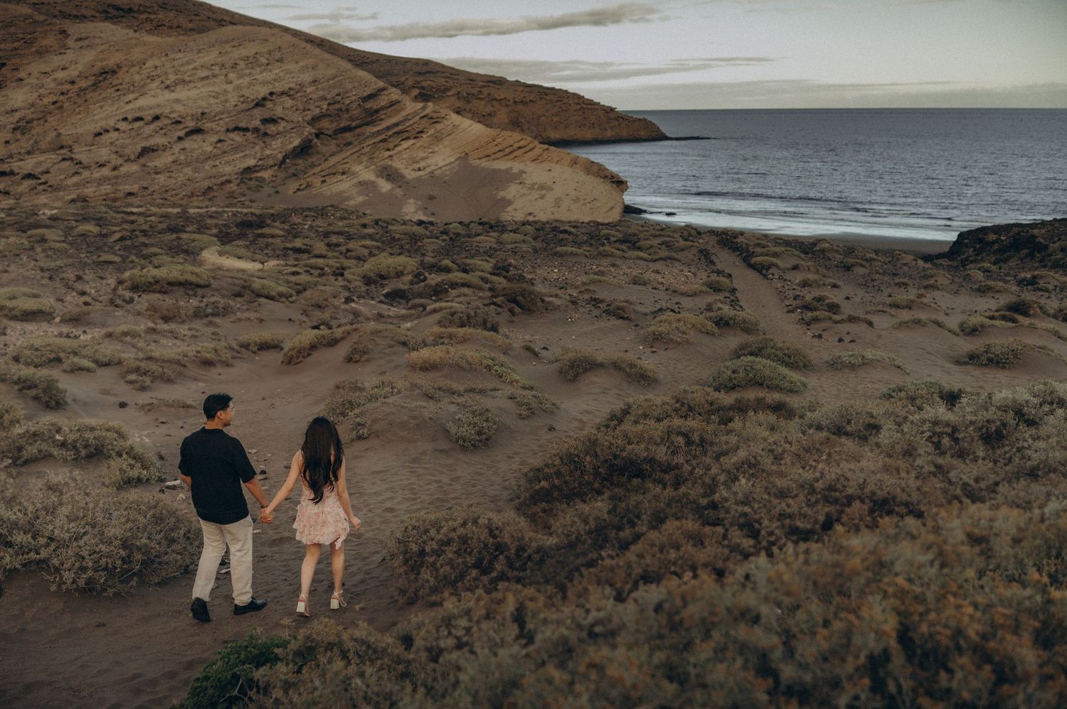 Couple holding hands overlooking a scenic coastal landscape with sandy hills and ocean views at dusk.