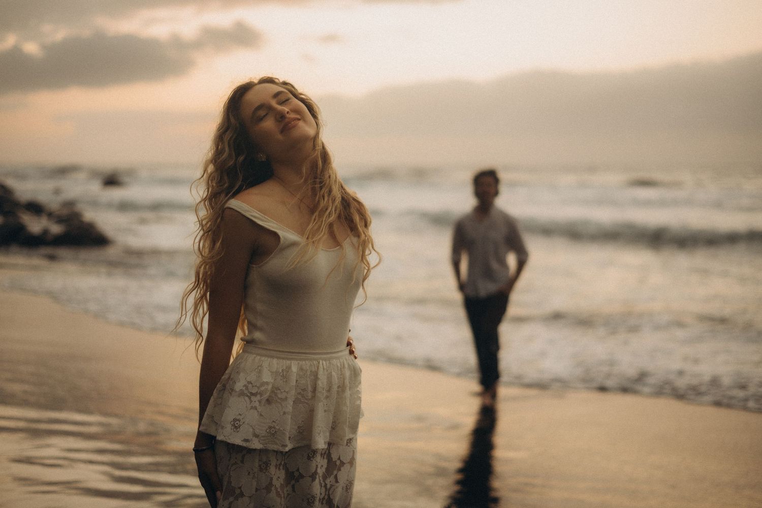 Woman with flowing hair enjoys sunset on beach while man walks toward her from ocean waves.
