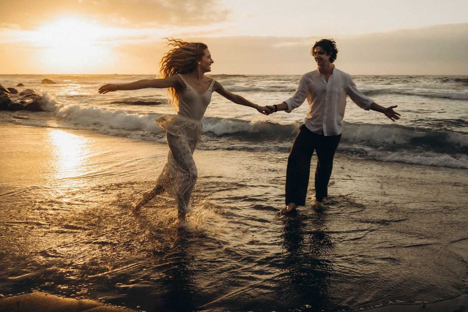 Couple holding hands dancing in ocean waves at golden sunset on beach.