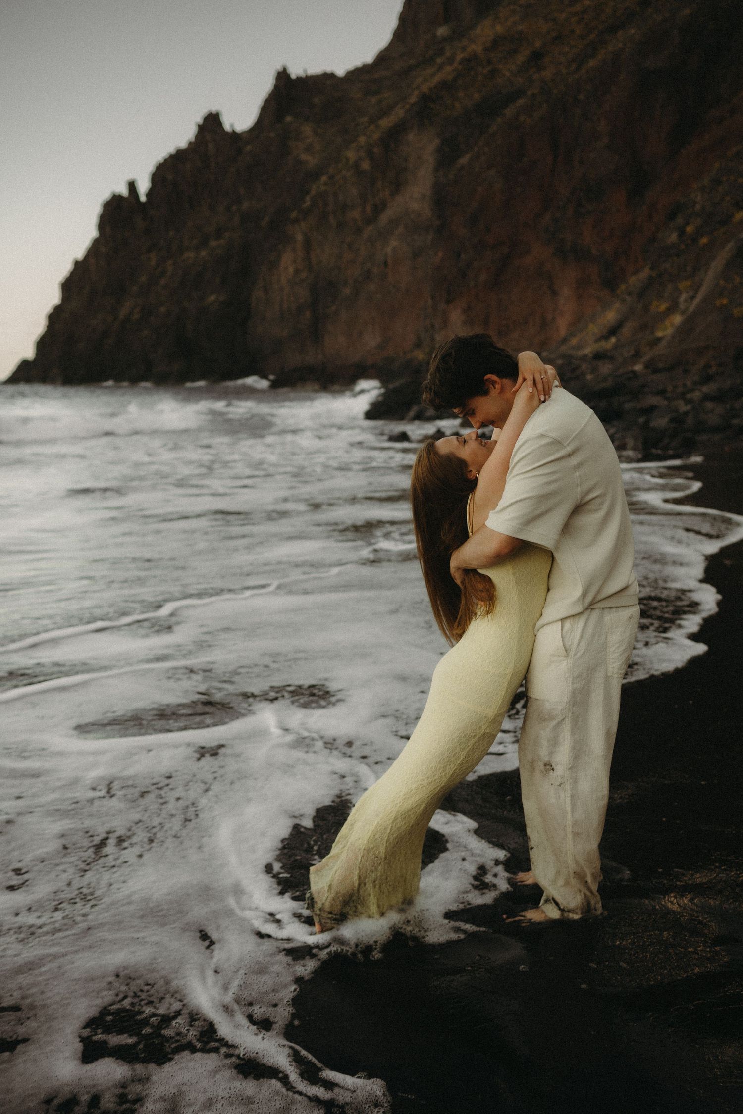 Couple kissing on a black sand beach with ocean waves and dramatic rocky cliffs in the background.
