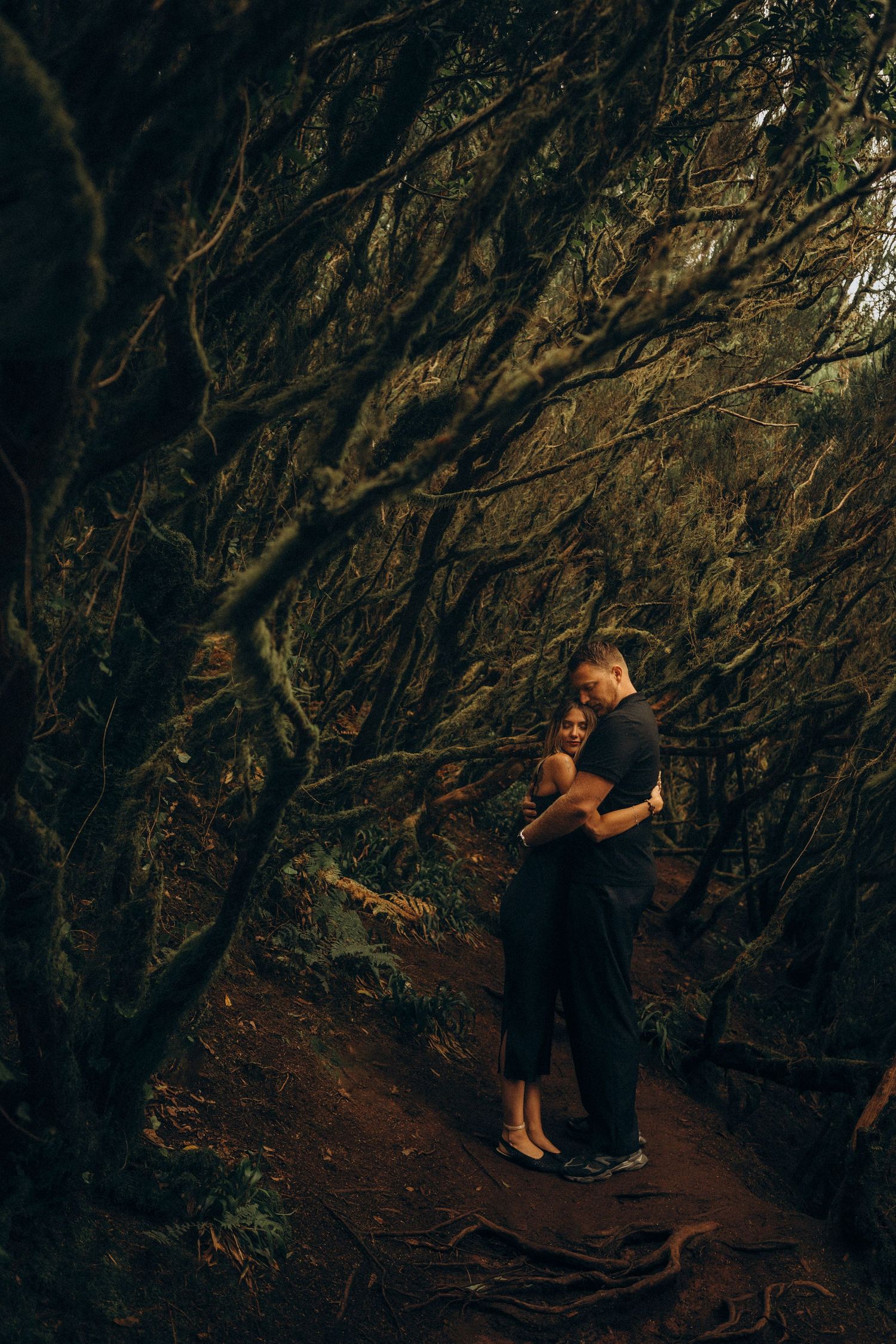 Couple embracing on a forest path surrounded by dark, moss-covered twisted trees in a romantic woodland setting.