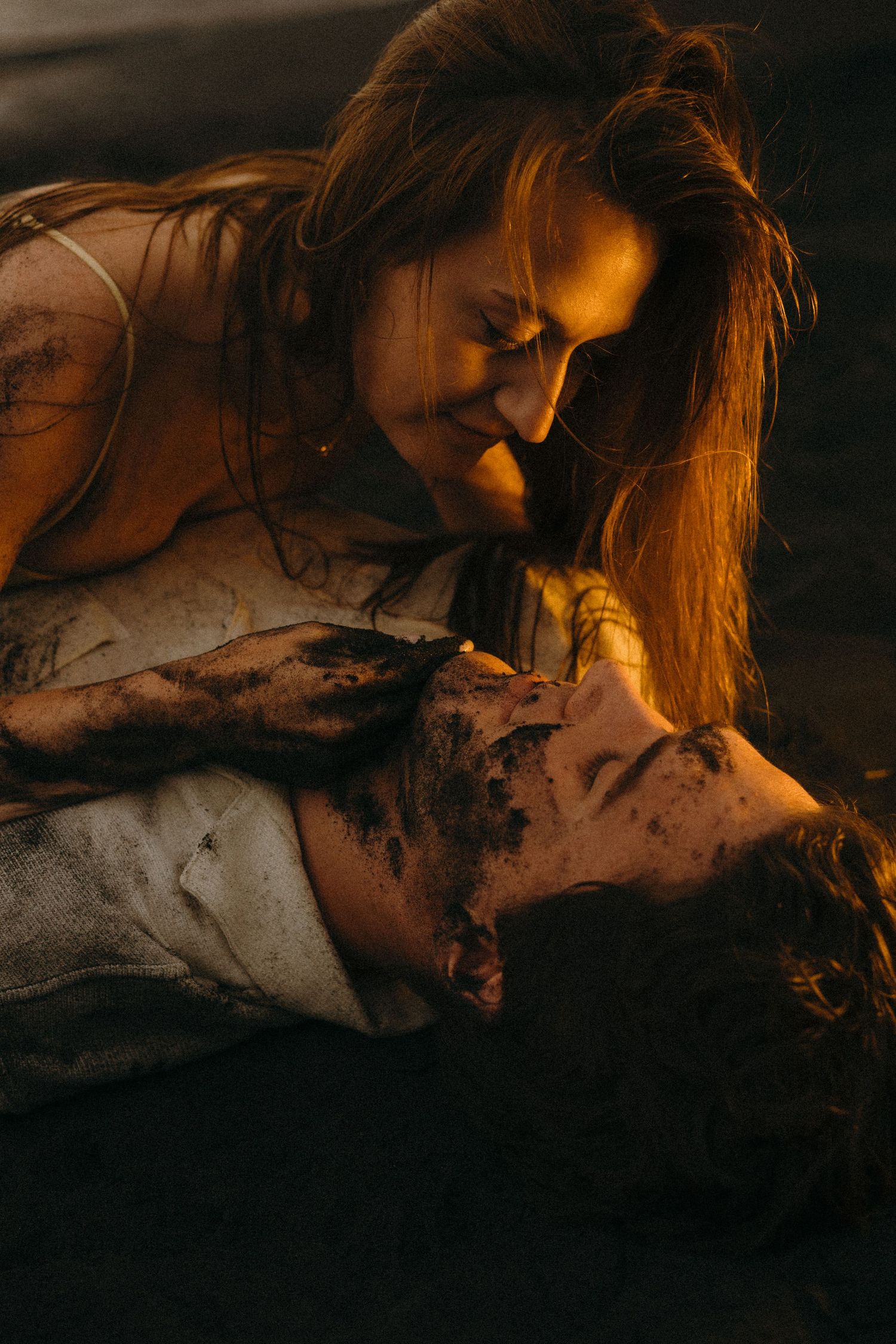 Two people covered in mud share an intimate moment, bathed in warm golden light outdoors.
