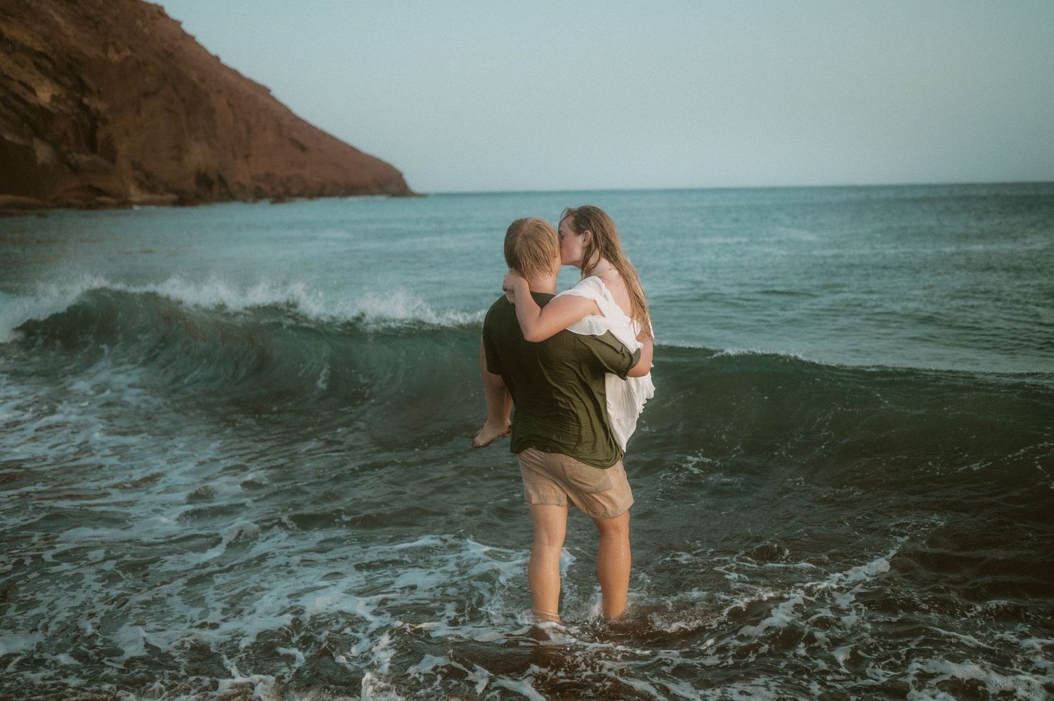 Man holding his partner in his arms as they jump together into the ocean waves, Playa Tejita, Tenerife couples photoshoot