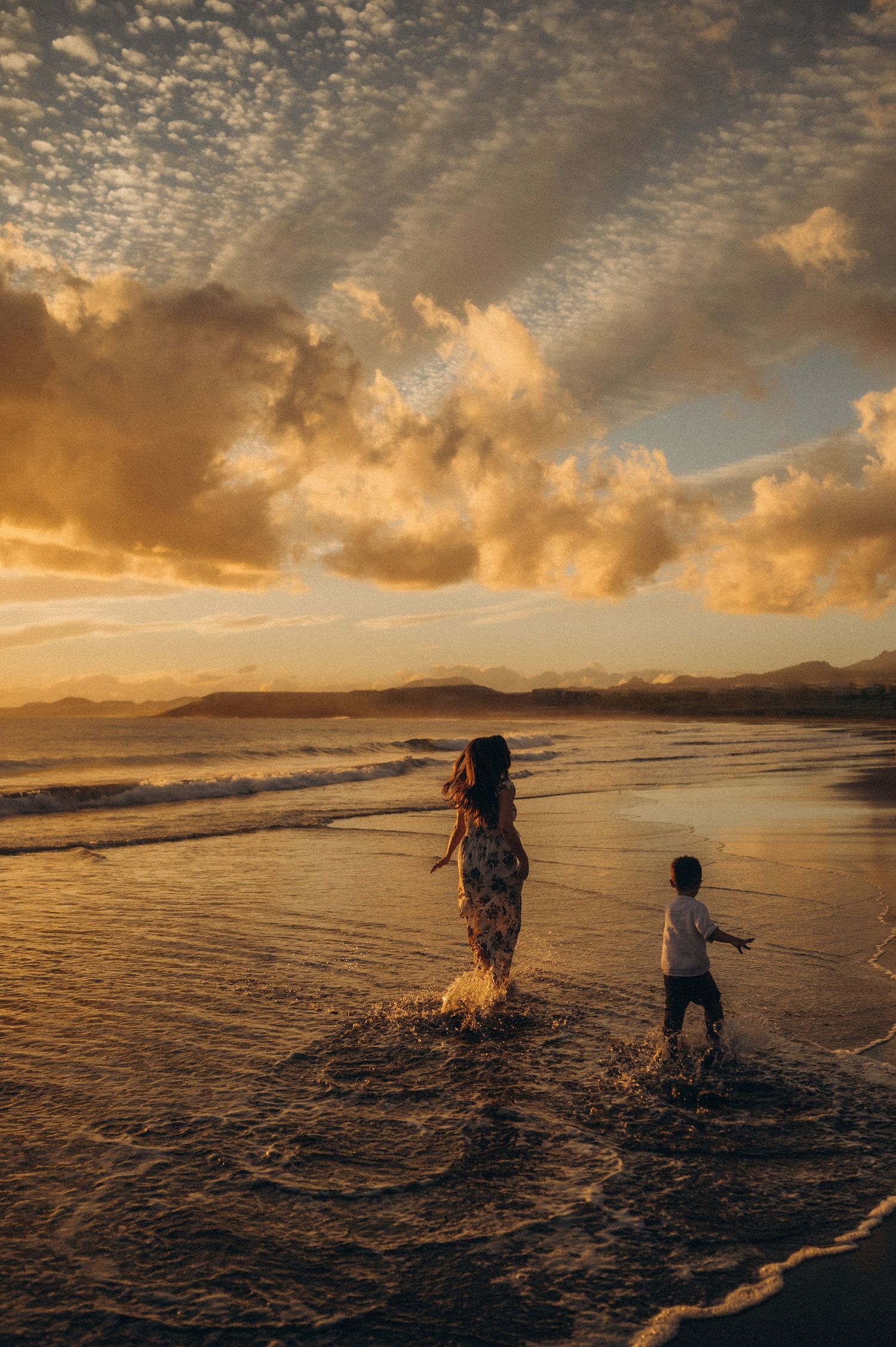 Silhouettes of people playing in ocean waves at sunset with dramatic golden clouds and mountains in background.